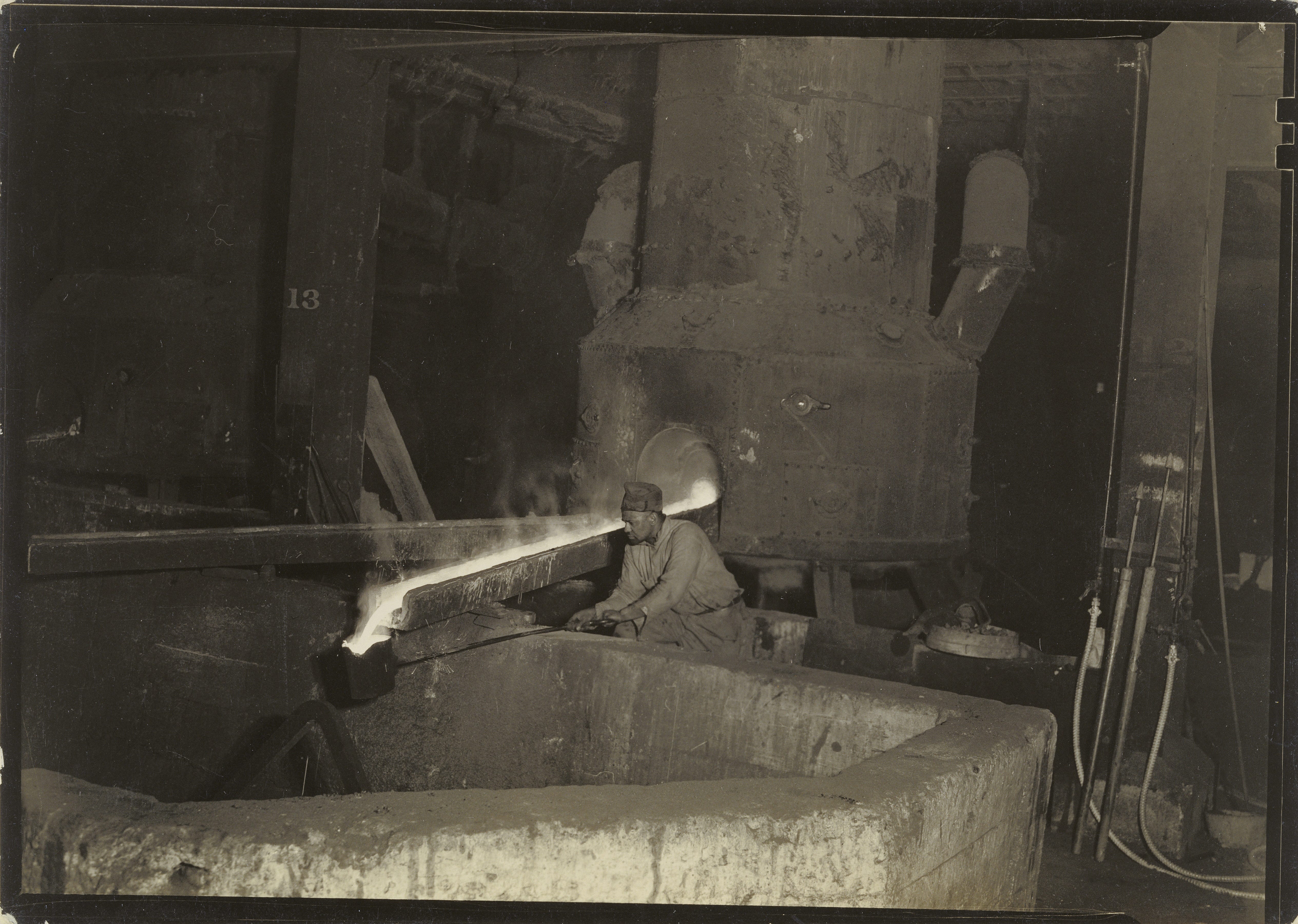 A worker hols a bucket on a long pole and catches liquid meta pouring from a large industrial device