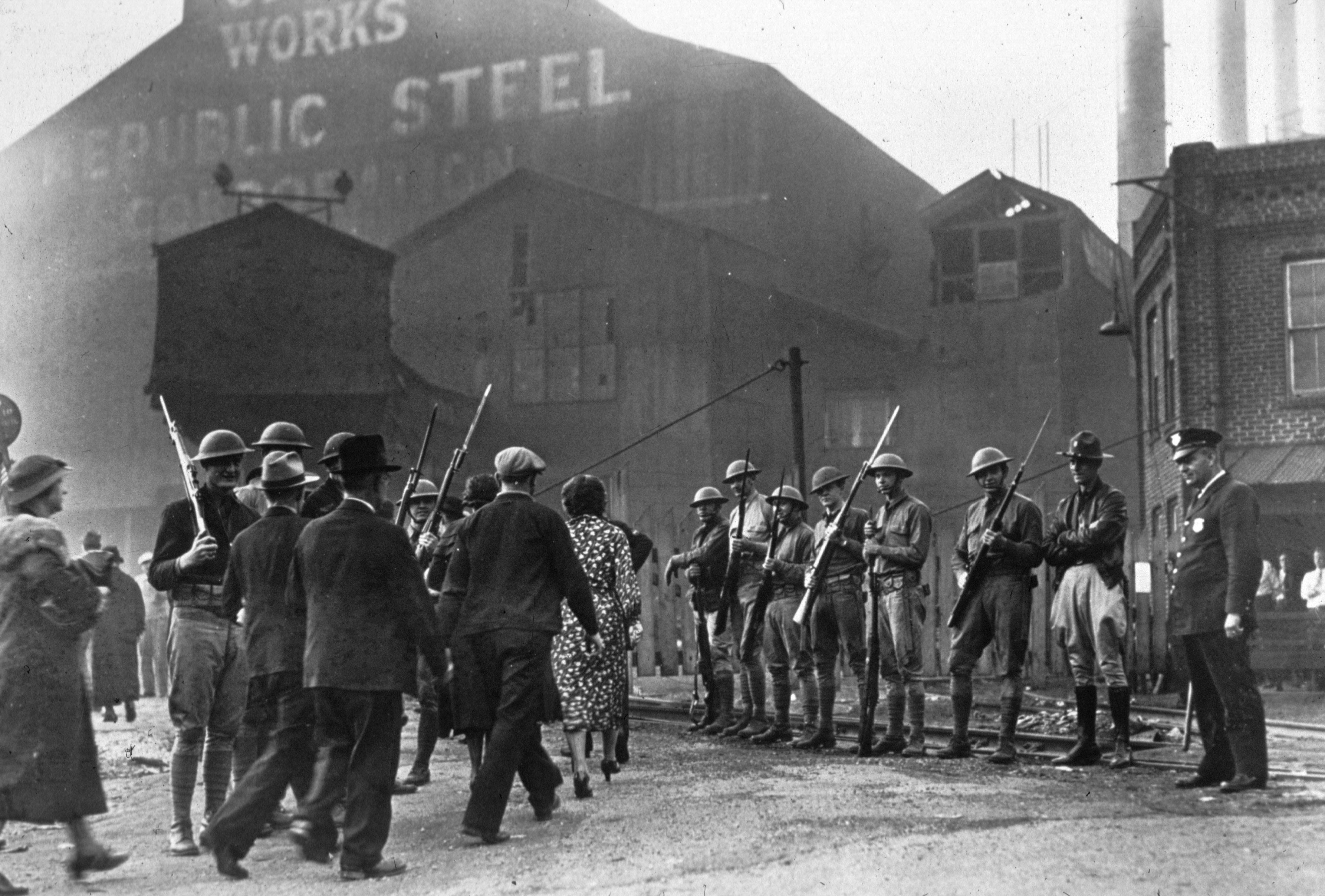 A crowd strikes outside a steel mill near men with guns wearing helmets