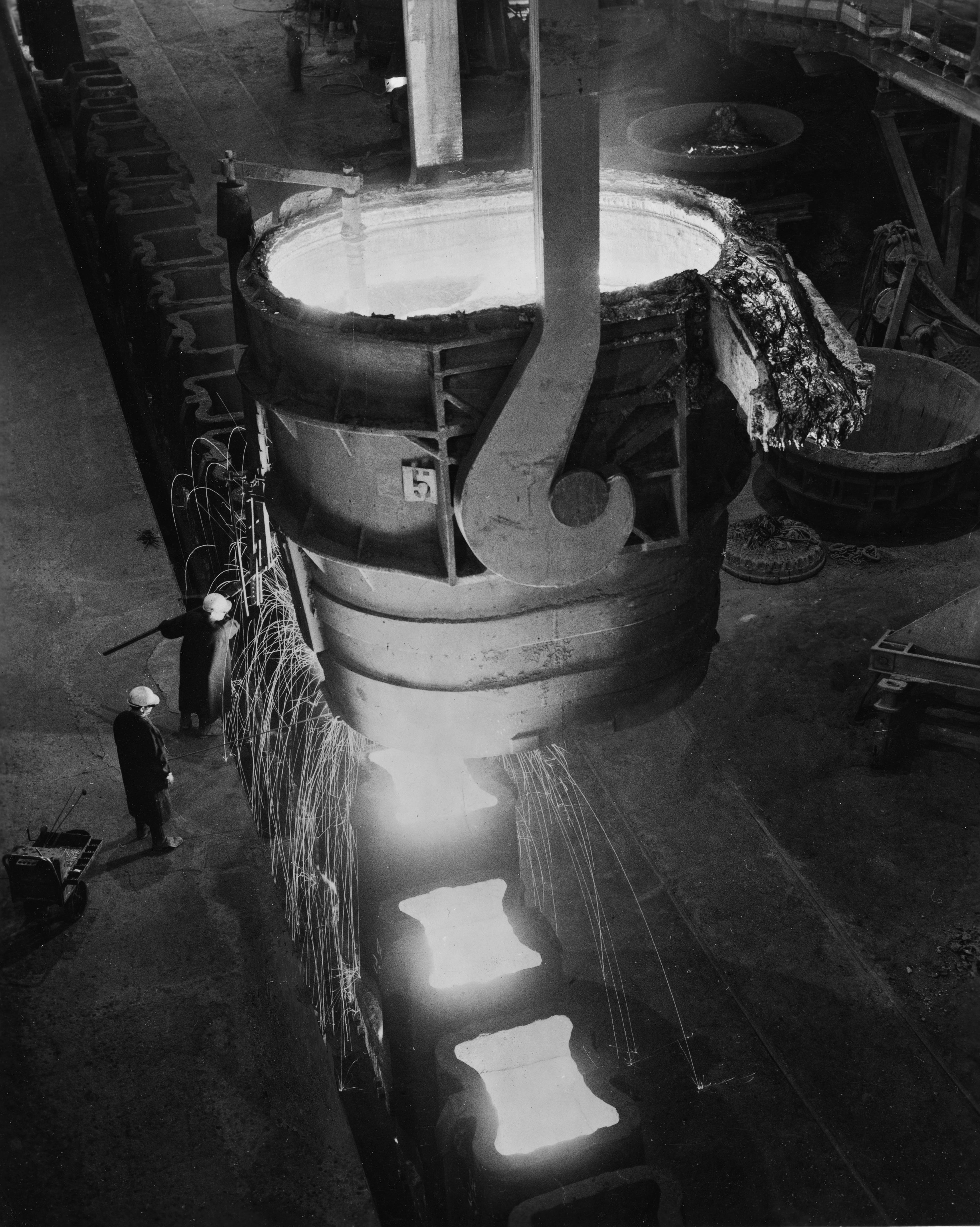 Two men stand below a large container of molten steel high above their heads