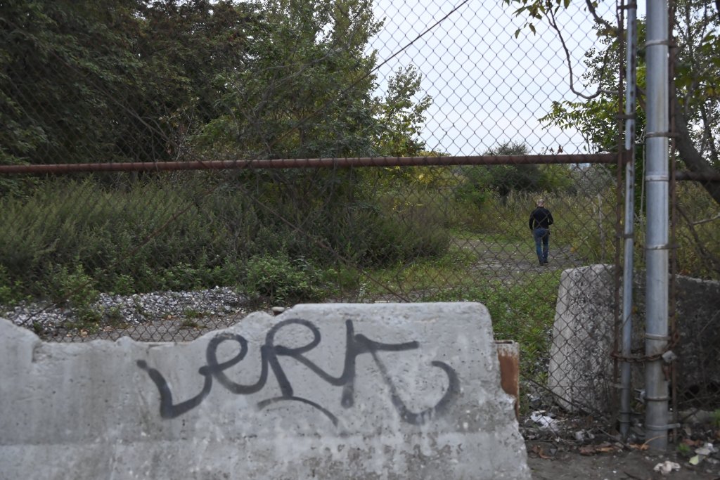A person walking away from the camera through an overgrown, fenced area with graffiti on a concrete barrier in the foreground.