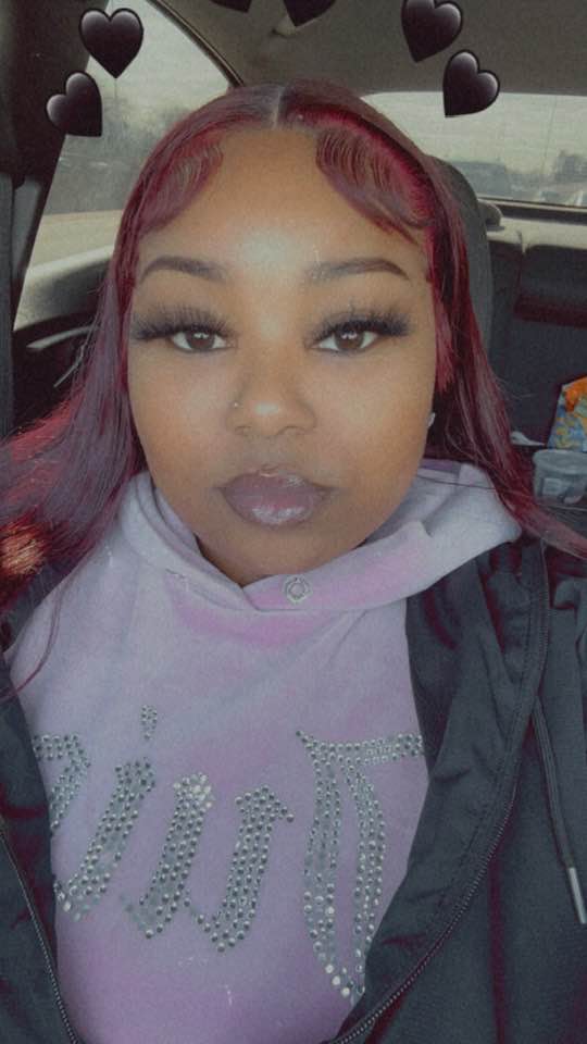 A woman in a car with reddish-purple hair, winged eyeliner, and a light purple hoodie.