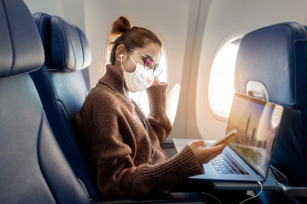 A woman wearing a face mask and glasses, with earphones, looking at her phone and laptop in an airplane.