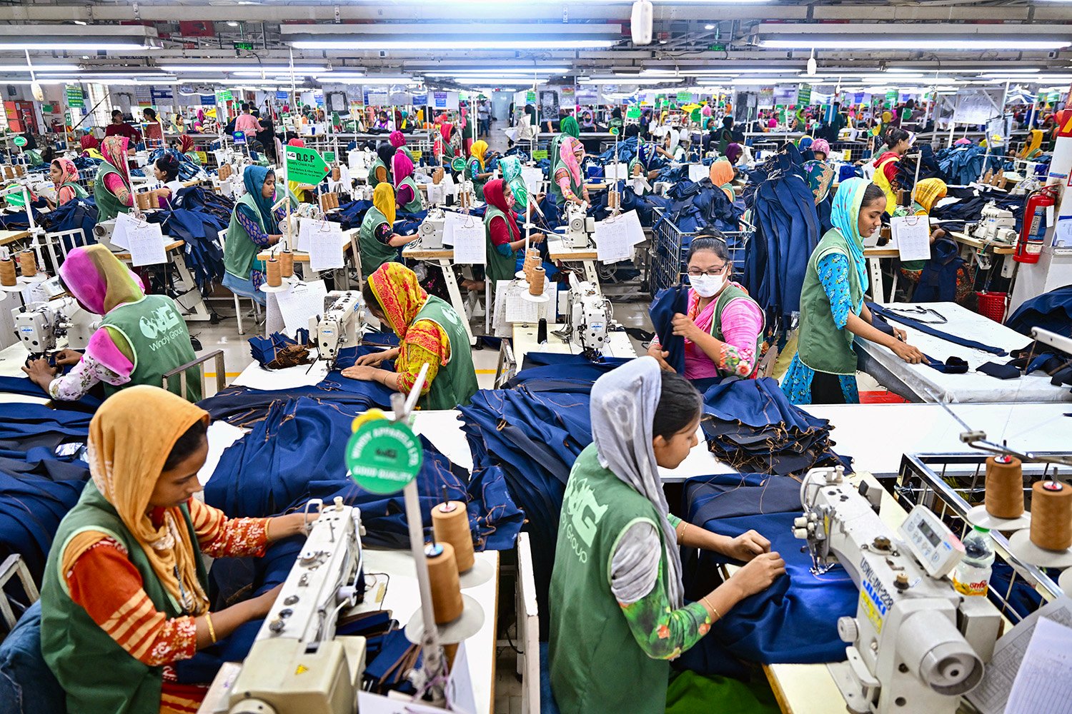 Dozens of women sit in rows at sewing machines as they work on a crowded textile factory floor. Piles of similar lengths of blue fabric take up many of the tables, but the women wear a rainbow of colors and patterns along with their matching green uniform vests.