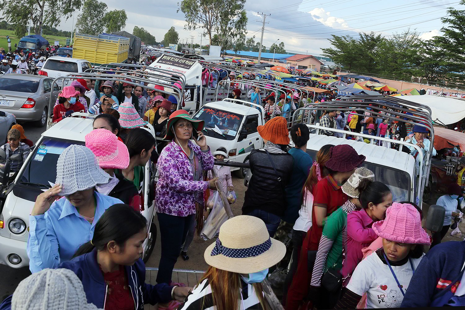 A crowd of workers and vehicles moves along a road outside of a factory. Most of the pedestrians milling out between vehicles are women, many wearing brimmed sun hats.