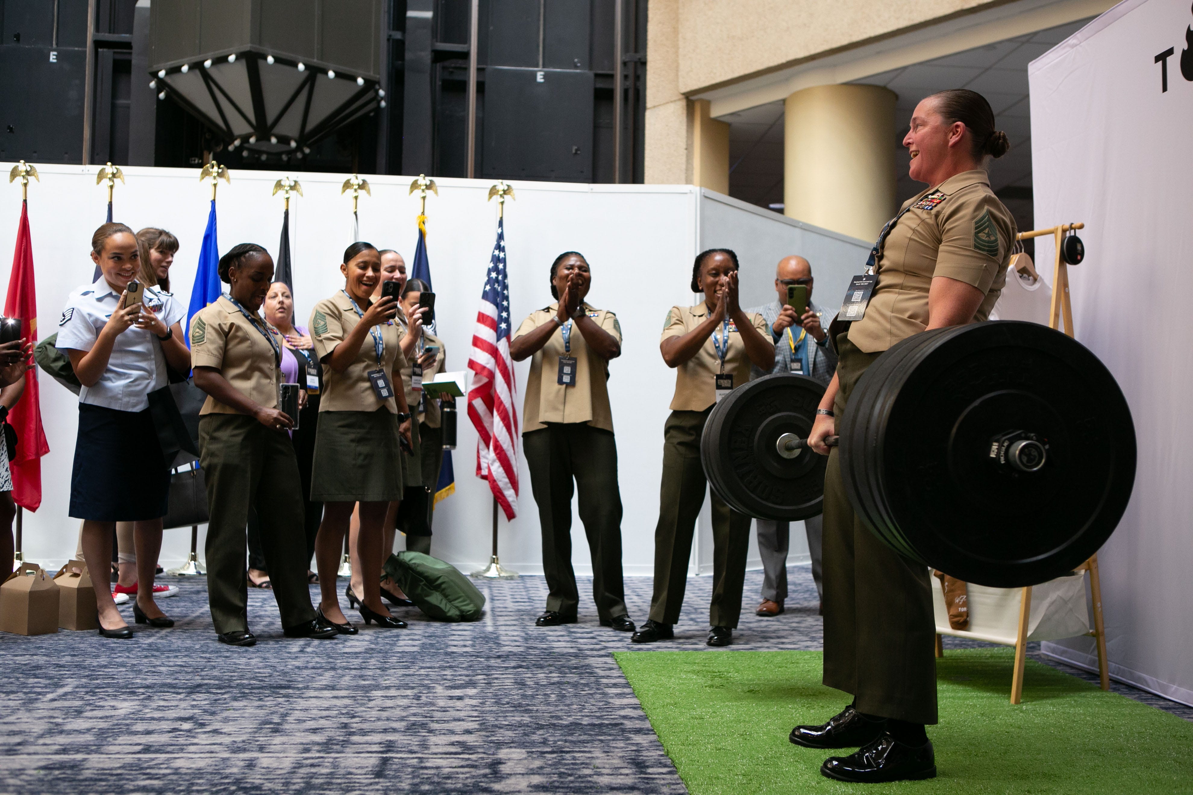 A Marine deadlifts while other service members cheer her on during the now-defunct Joint Women's Leadership Symposium.
