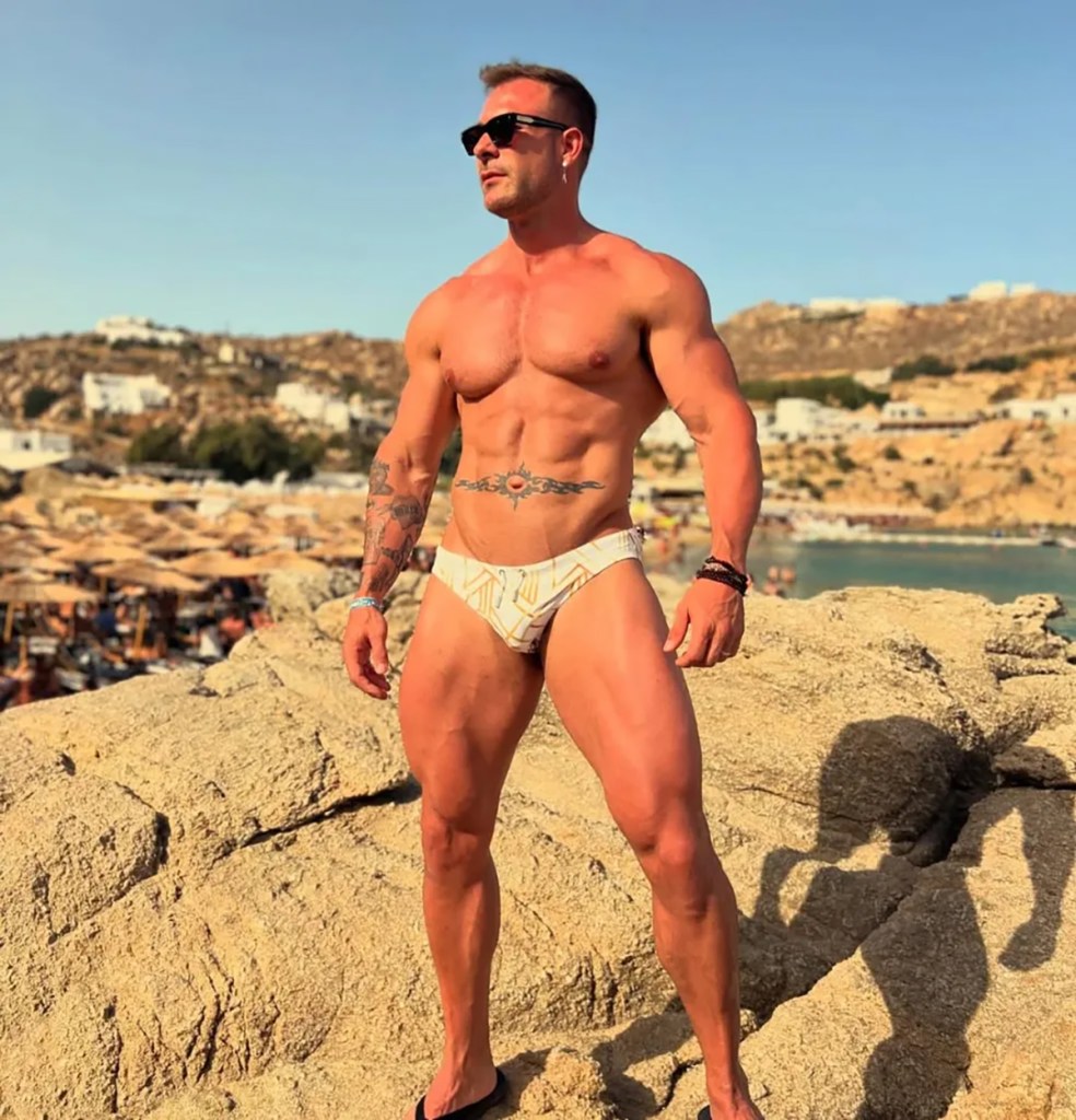 Justin Heath Smith shirtless, posing in white swimwear and sunglasses on a rocky beach.