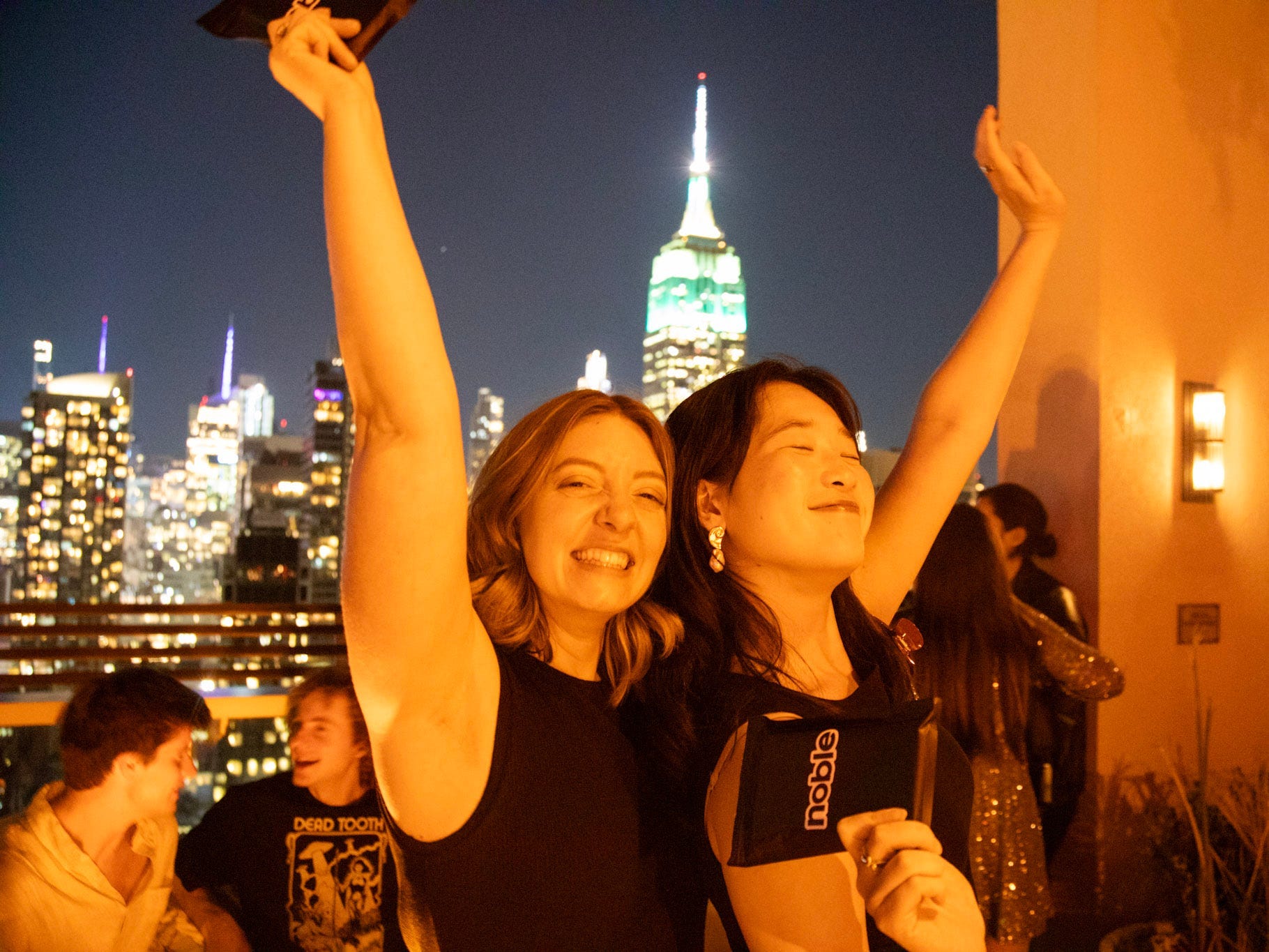 Two women raise their arms overhead in merriment at a rooftop party with the Empire State Building piercing the skyline behind them.