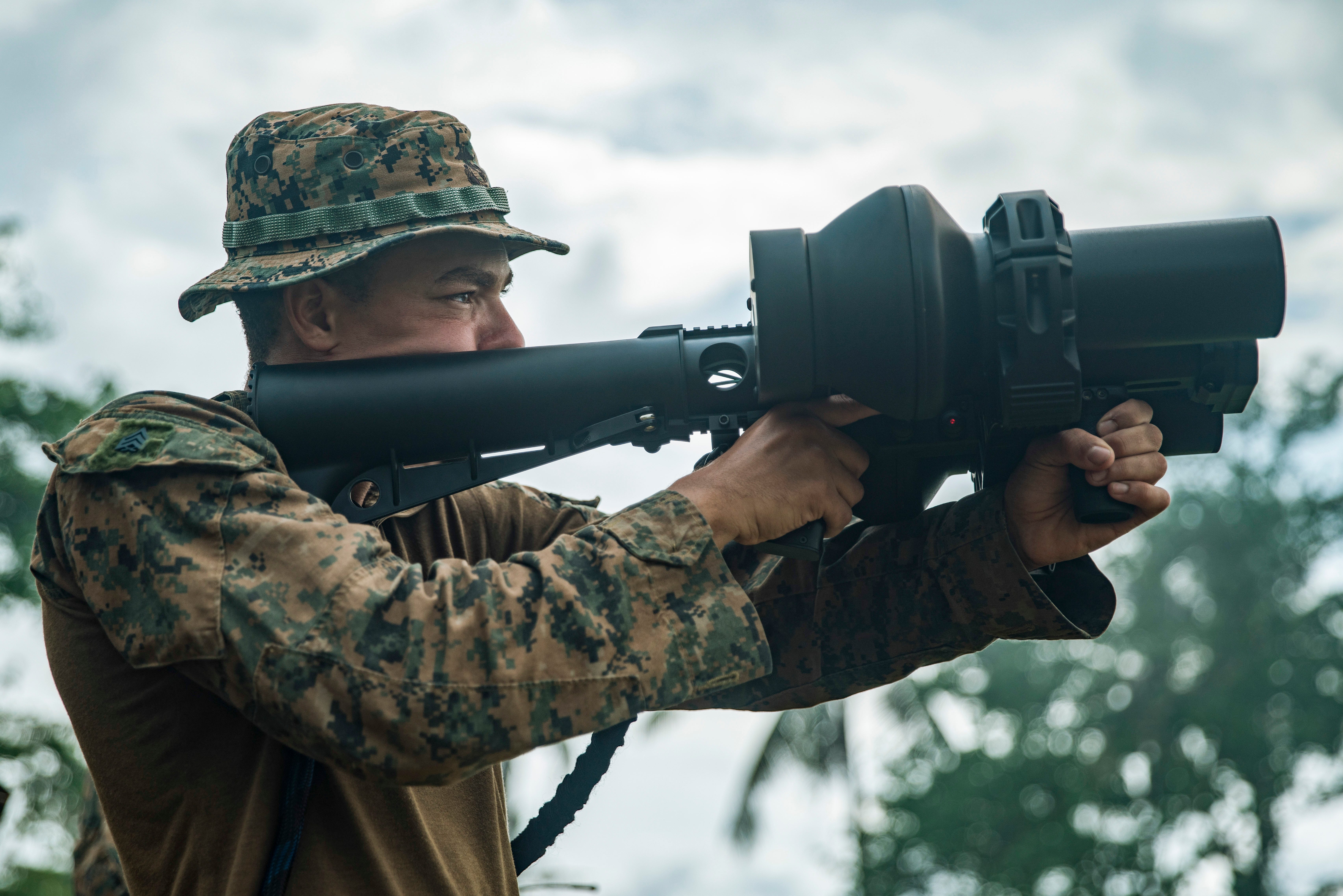 A man wearing camouflage holds a counter-drone weapon on his shoulder in front of his eyes like a rifle. There are trees and an overcast sky in the background.