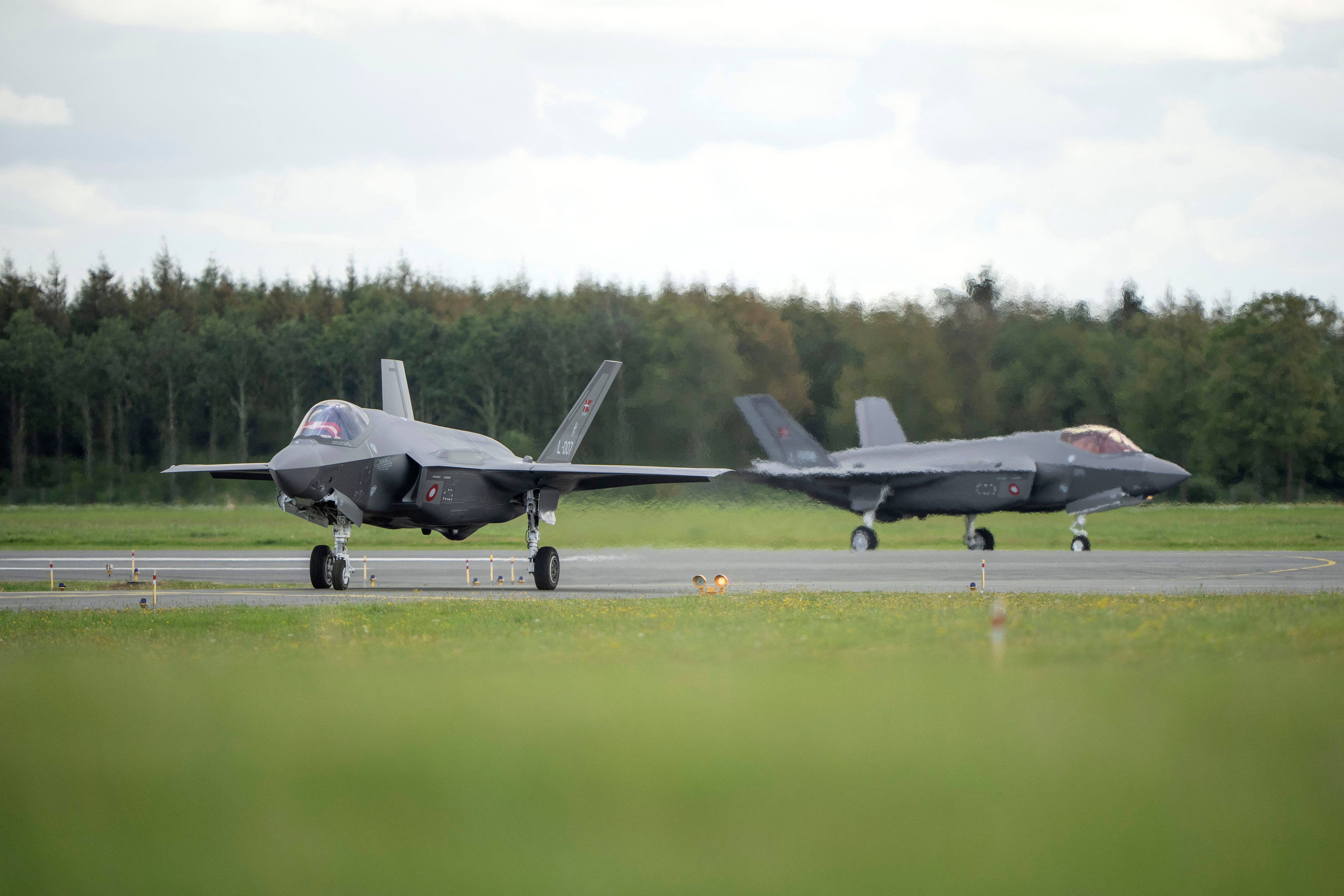 Two grey fighter jets on a runway with grass in the foreground and trees behind them