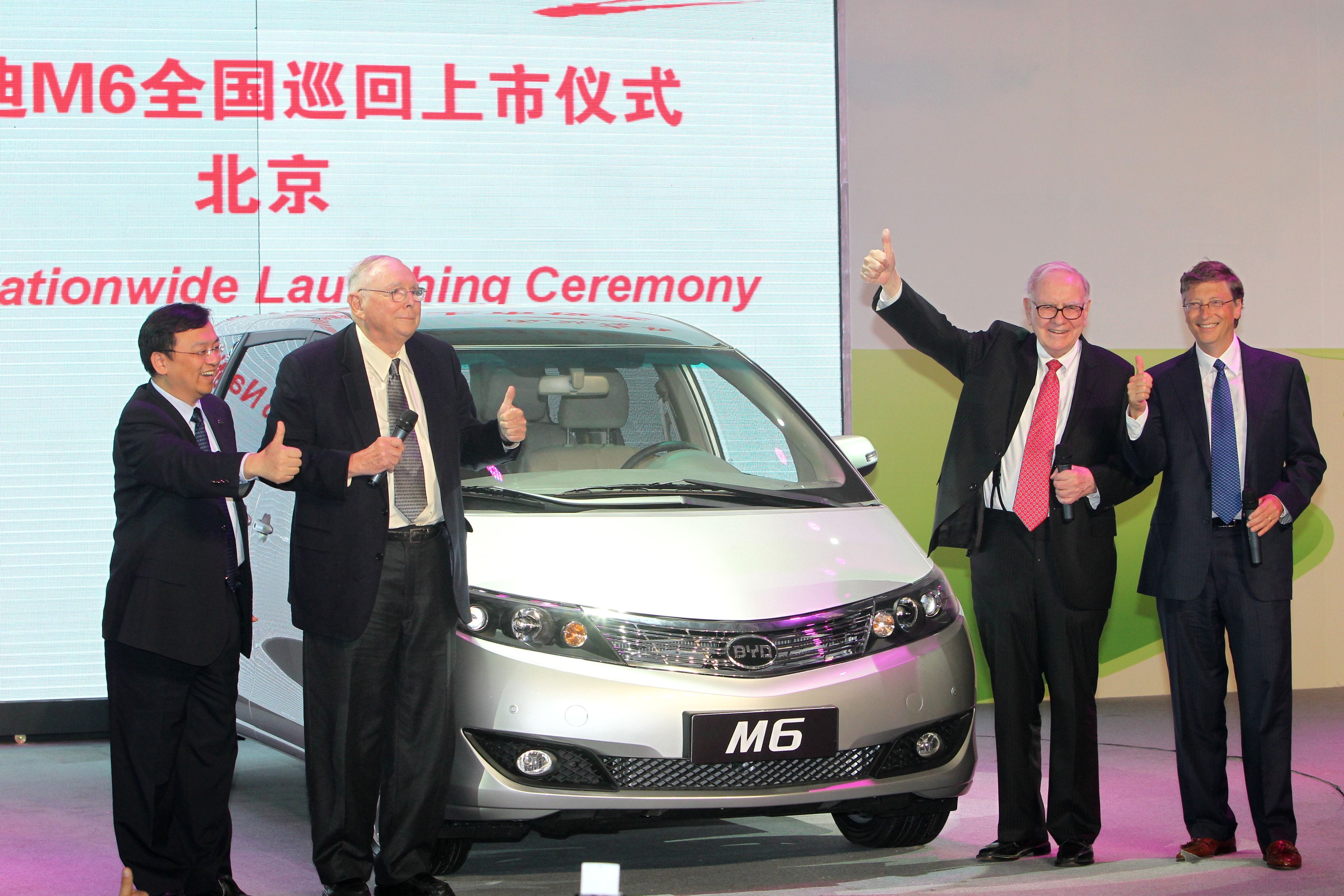 Wang Chuanfu, Charlie Munger, Warren Buffett, and Bill Gates standing next to a BYD M6 and each giving a thumbs-up.