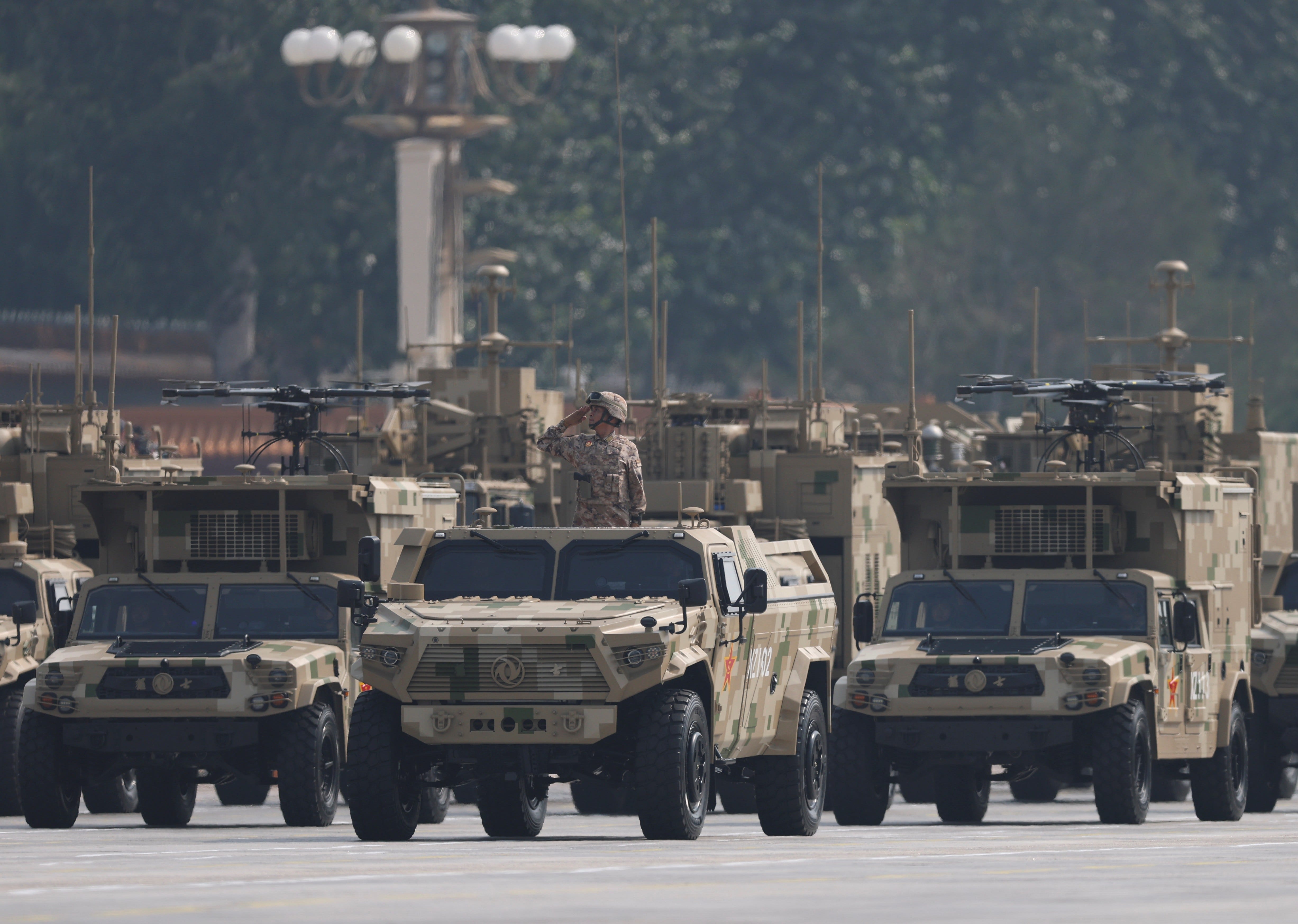 Chinese military trucks are lined up on a road. A soldier stands in one. The two on other side of him have drones on top.