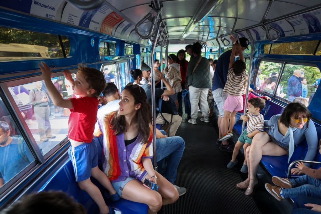 People riding a blue bus with various advertisements on the ceiling, including one that reads 