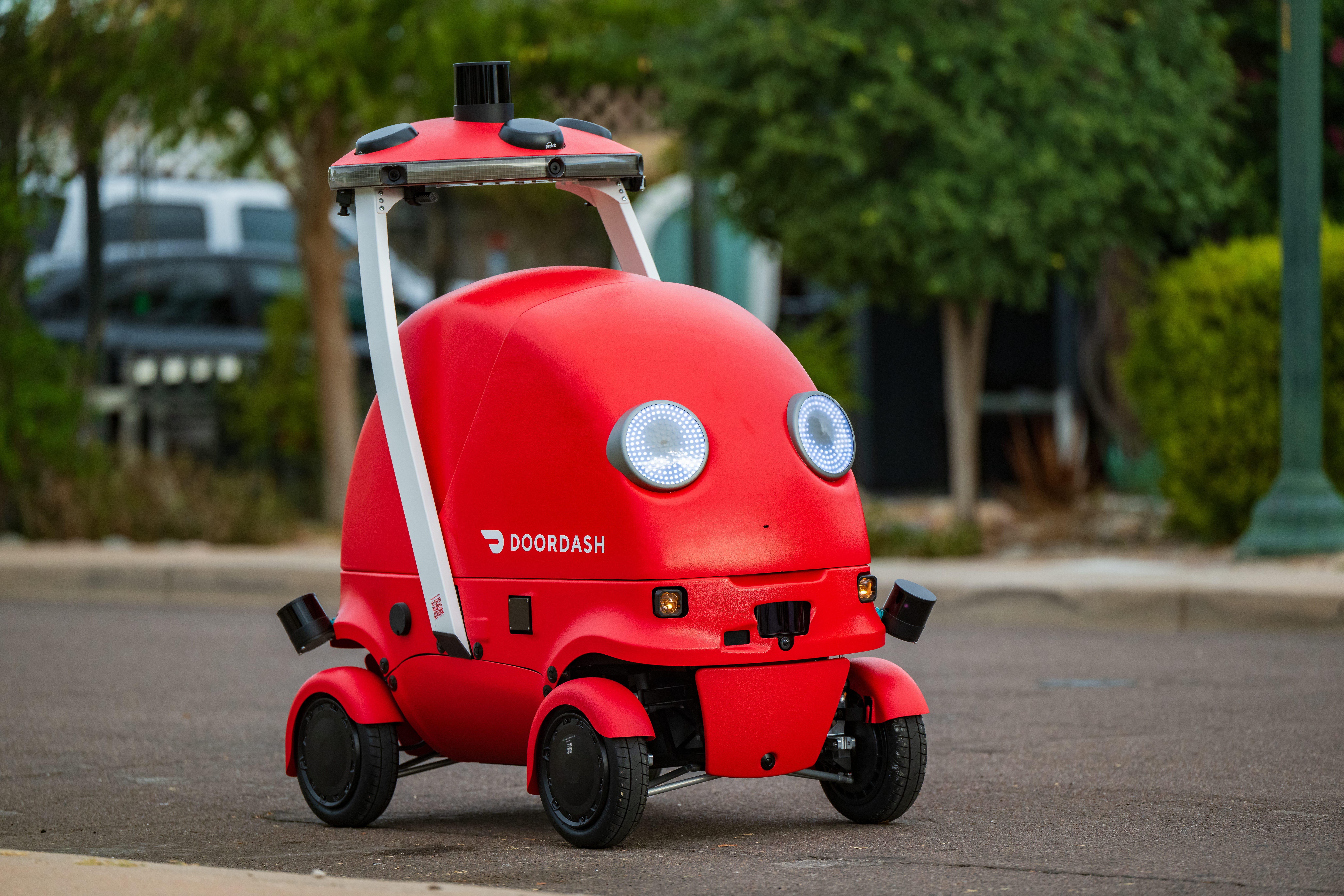 Dot, DoorDash's self-driving delivery robot, with a red outer shell, four black wheels, and two LED lights on its front that look like eyes, drives along a road.