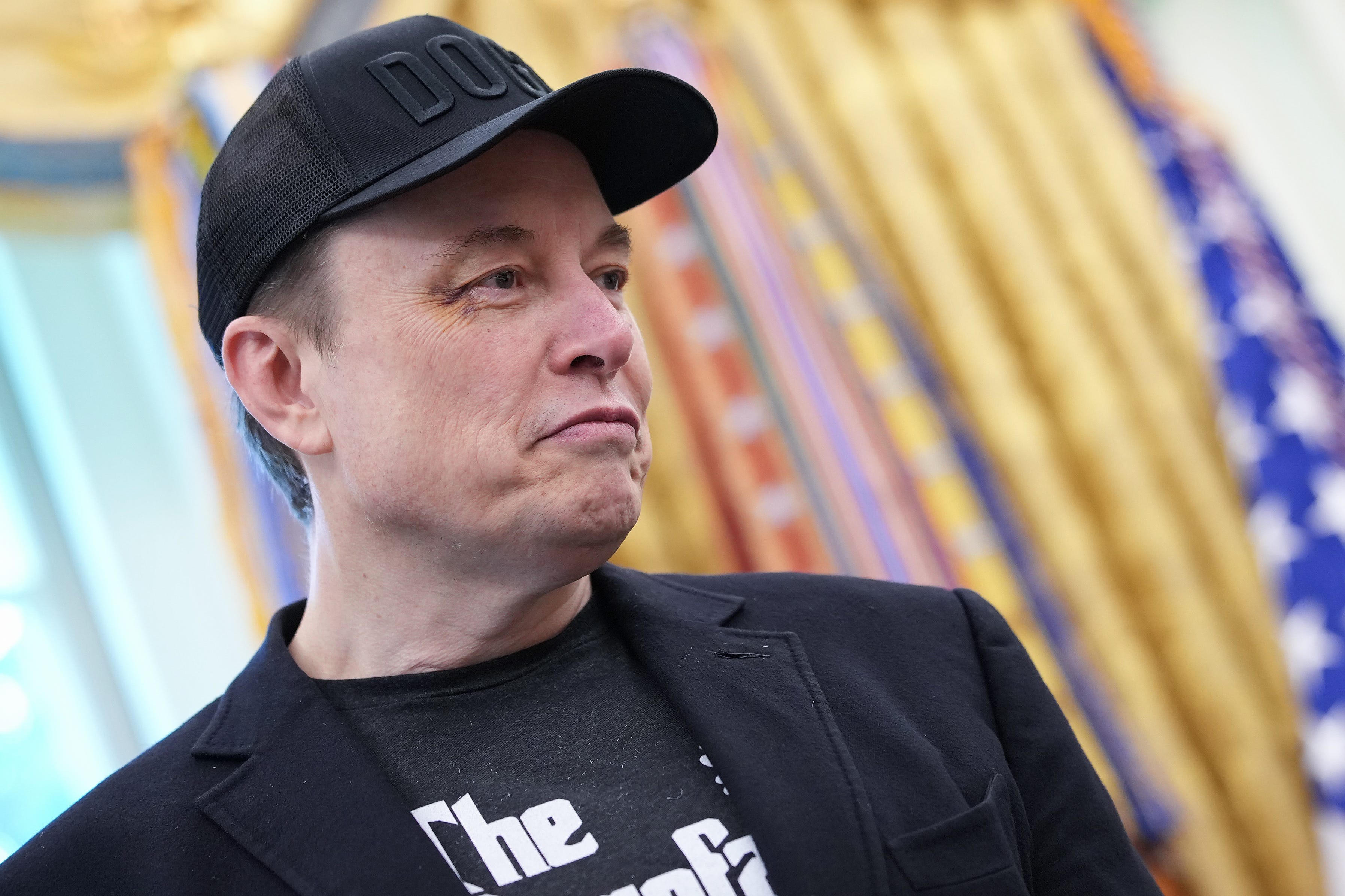 Elon Musk is wearing a DOGE cap and looking downwards while in the Oval Office at the White House.