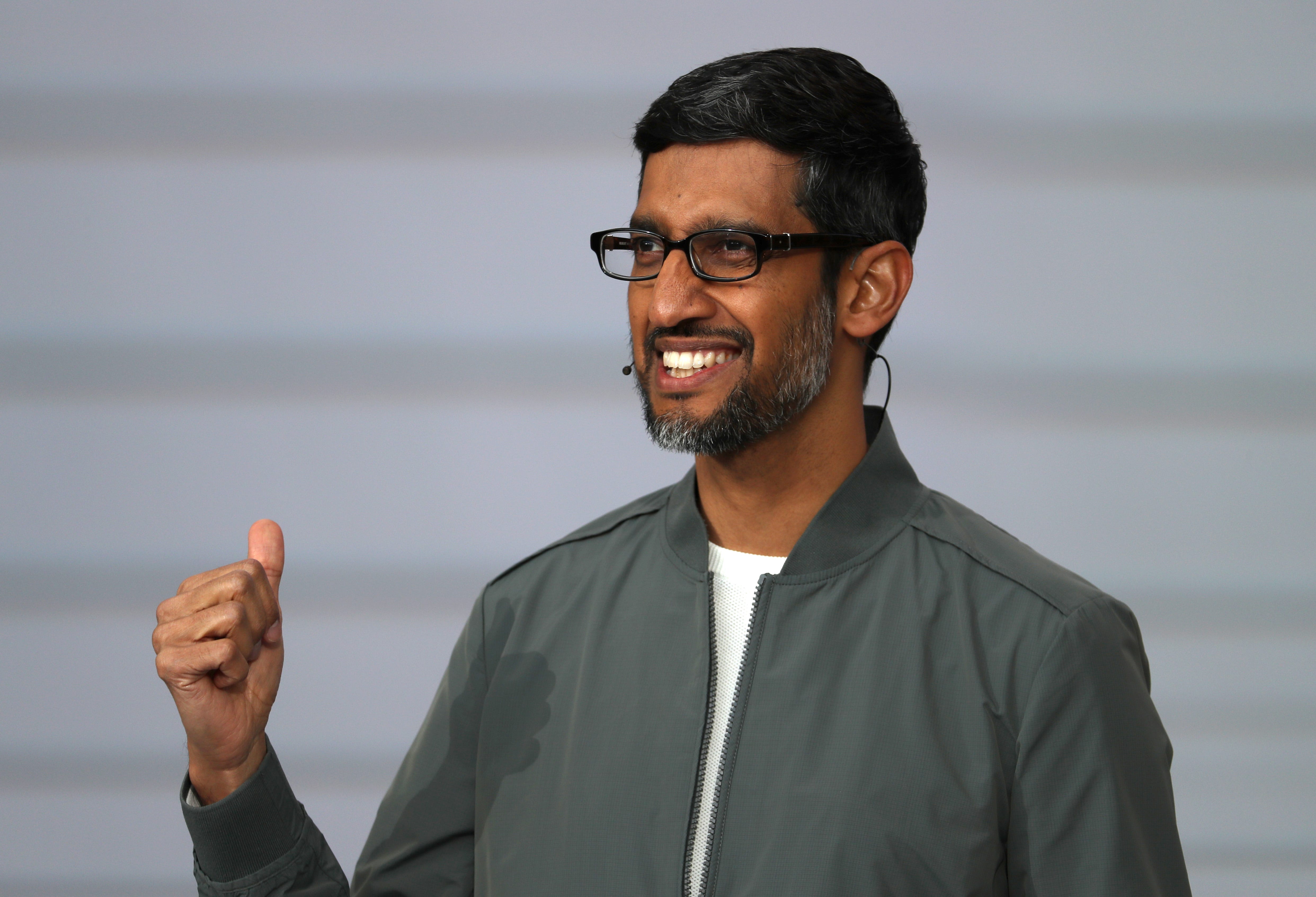 Sundar Pichai wears a grey jacket over a white t-shirt and smiles on stage.