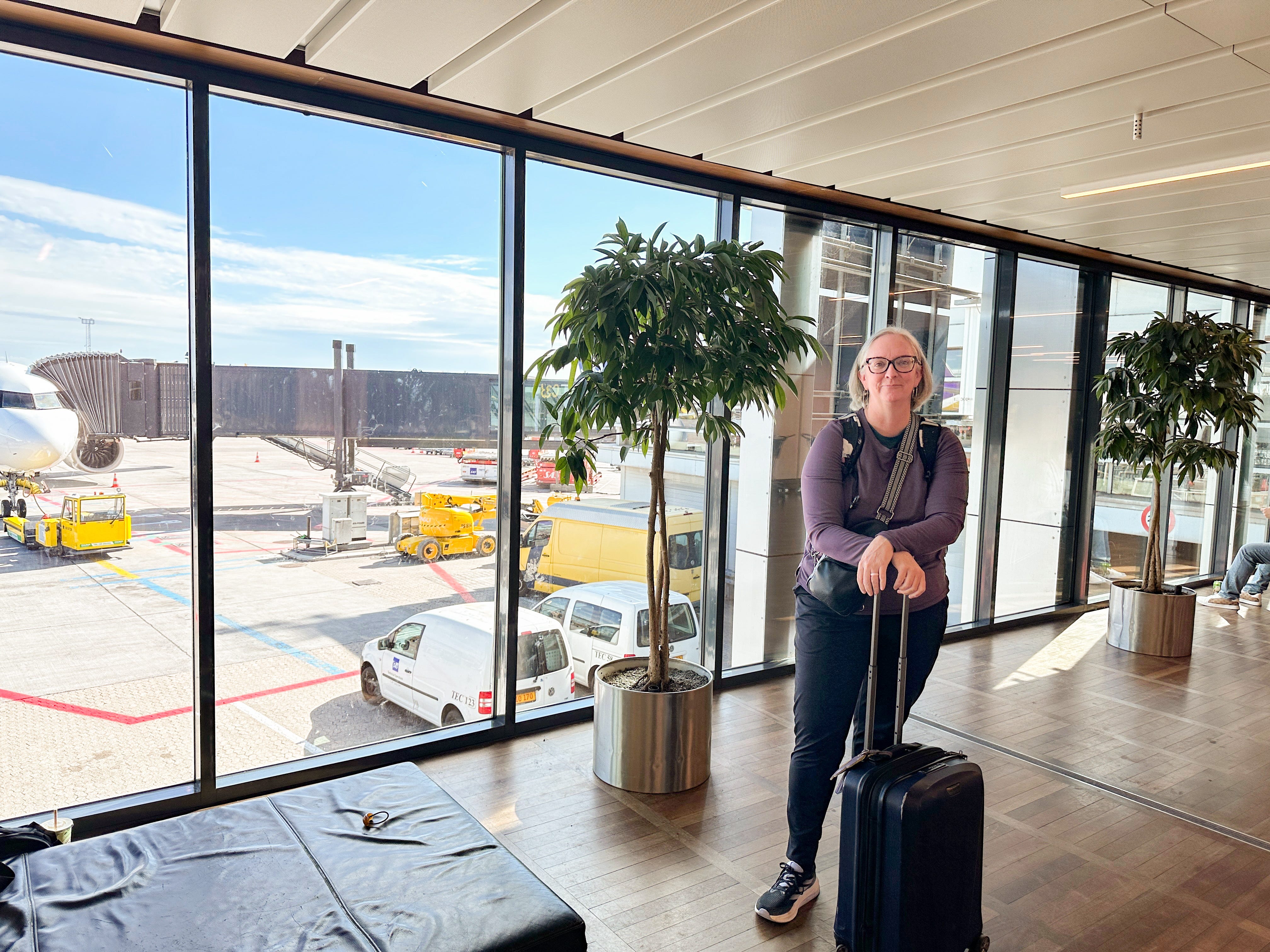 Kate stands in front of a window at the airport with a carry-on suitcase.