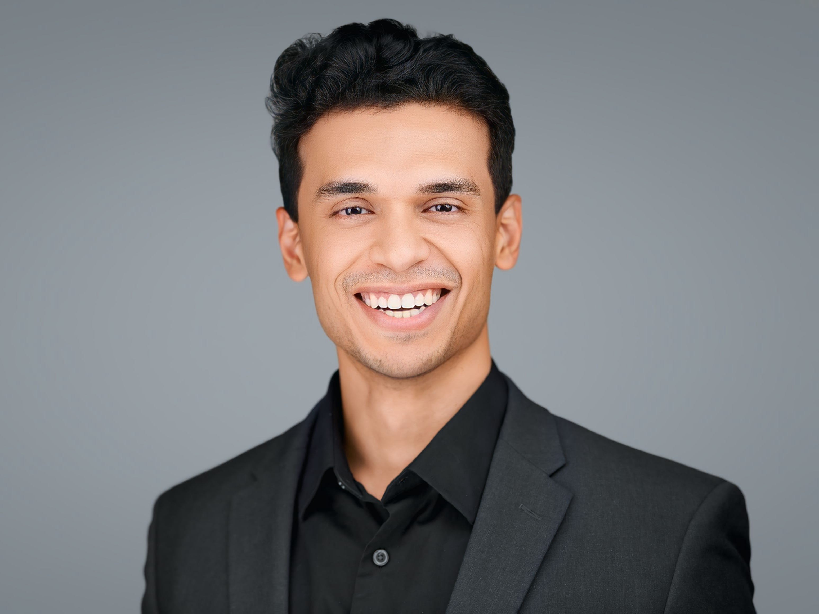 a man smiling and posing for a headshot in a black suit in front of a gray background.
