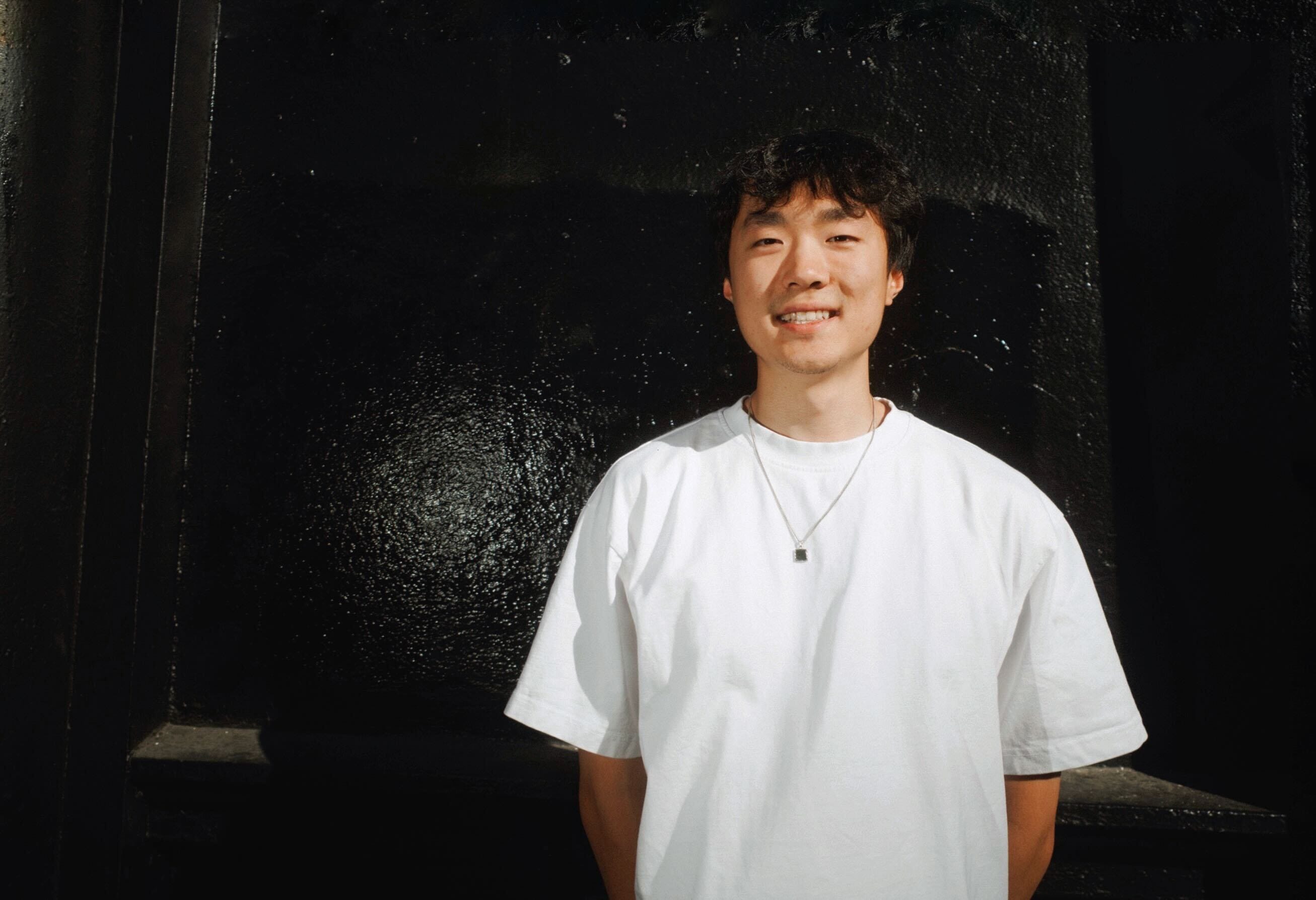 Lambert Liu is wearing a white T-shirt and posing in front of a black-colored wall.