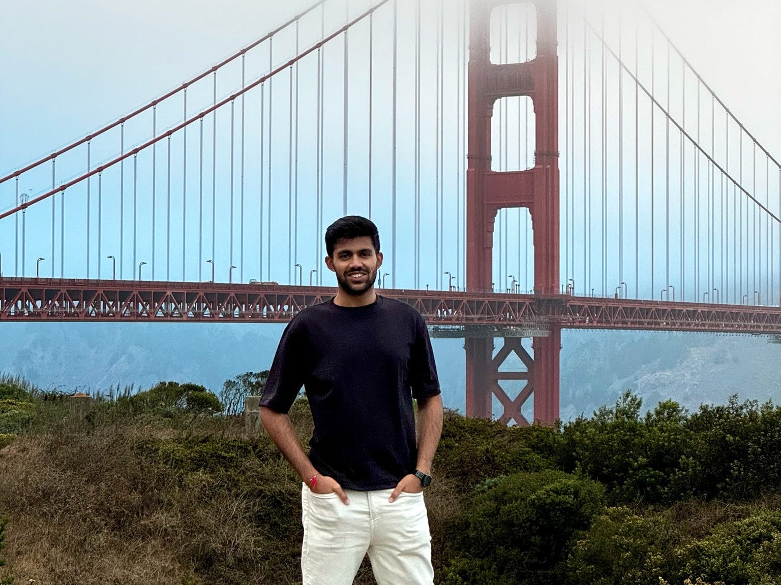 A  man stands smiling in front of the Golden Gate Bridge on a foggy day, with the bridge's top partially hidden in the mist. They are wearing a dark t-shirt, white pants, and white sneakers, posing with hands in pockets.