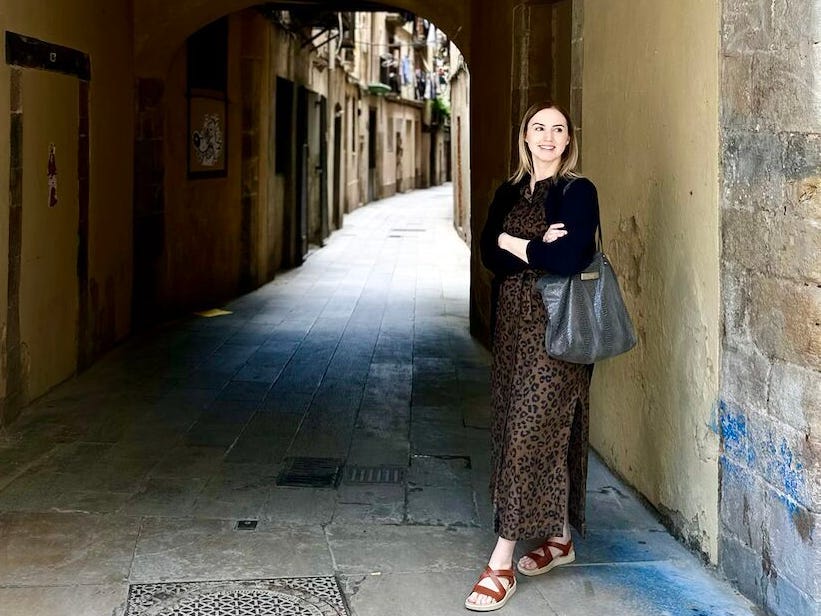 Woman standing under a stone archway in a narrow alley, smiling with arms crossed.