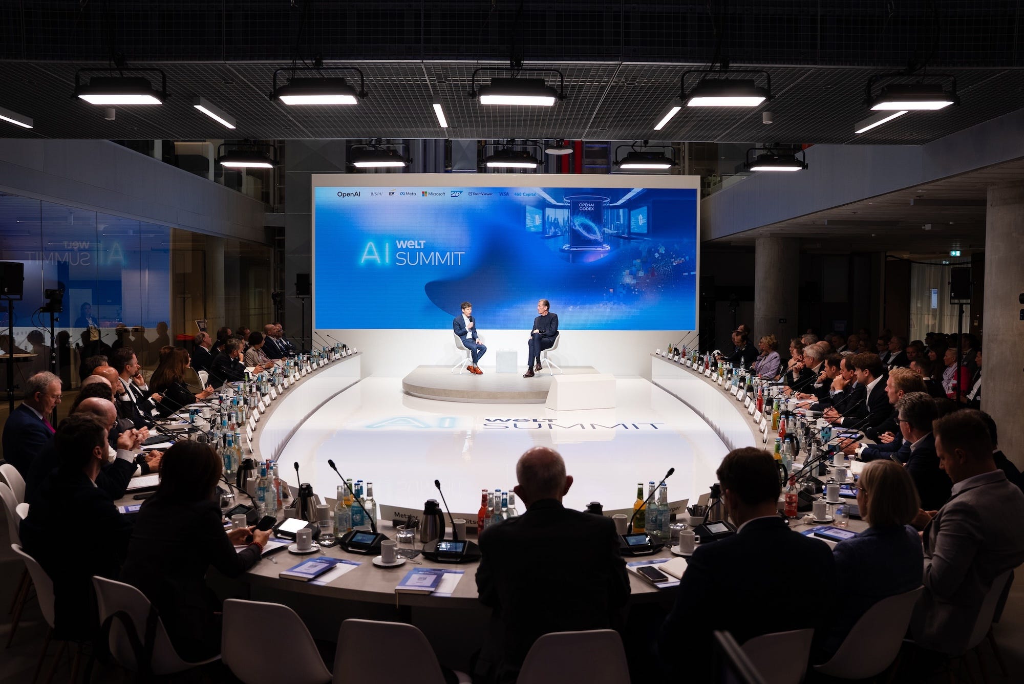 Sam Altman and Mathias Döpfner talk on a brightly lit stage surrounded by professionals.