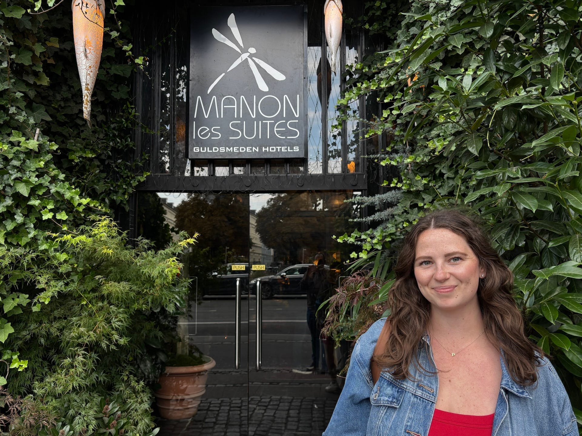 A woman with brown hair smiles in front of a sign that says "Manon Les Suites."