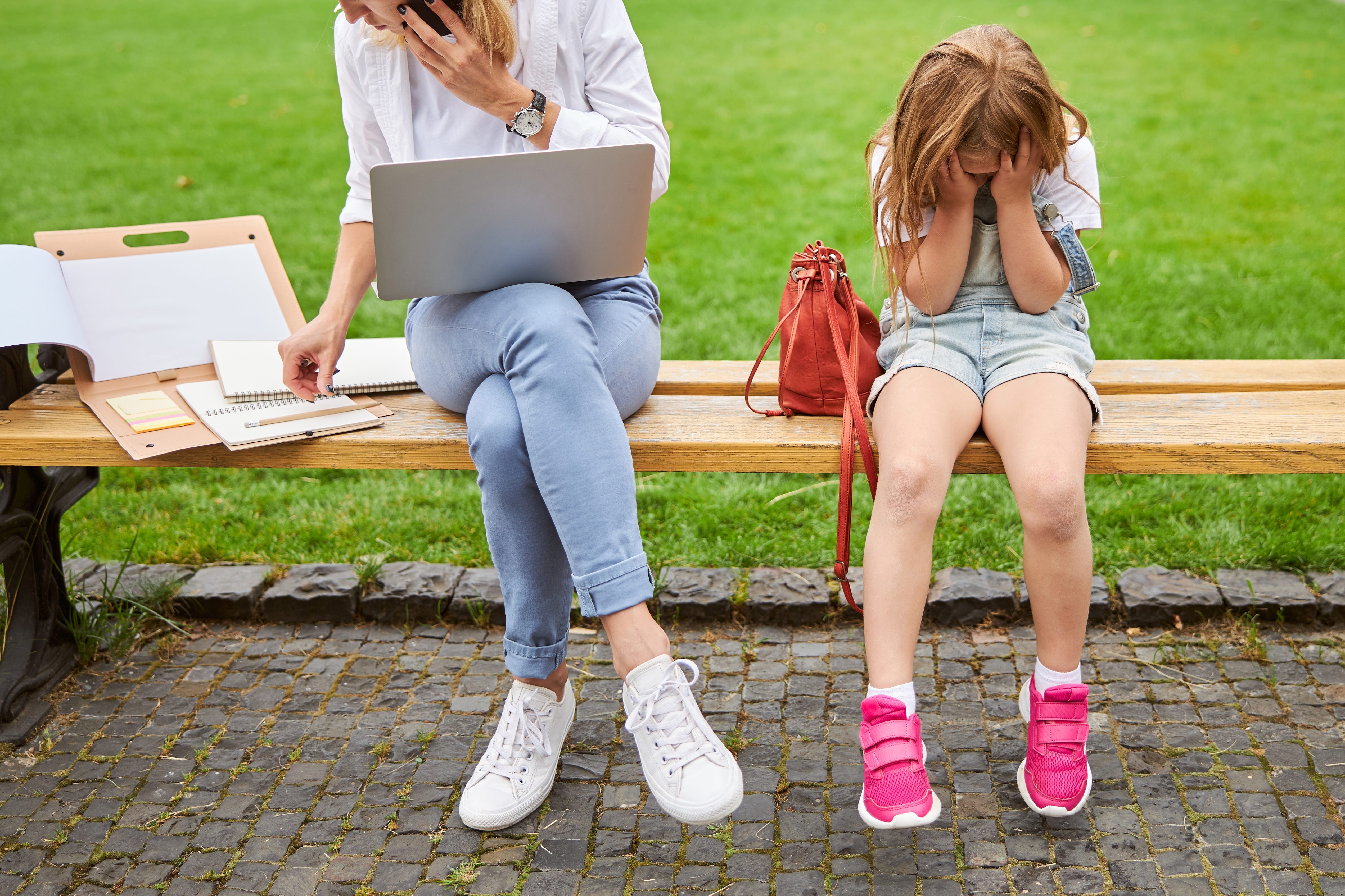 Busy mother working in park on wooden bench while a young girl sits by her side