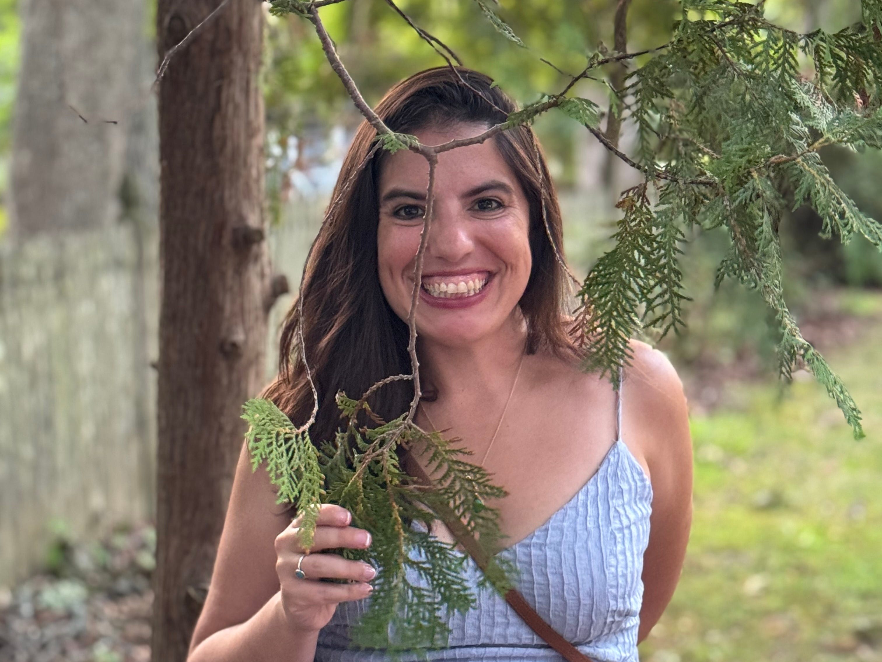 A woman in a light blue sundress stands outdoors smiling at the camera. She holds a leafy tree branch in front of her, with more greenery surrounding her in the background.