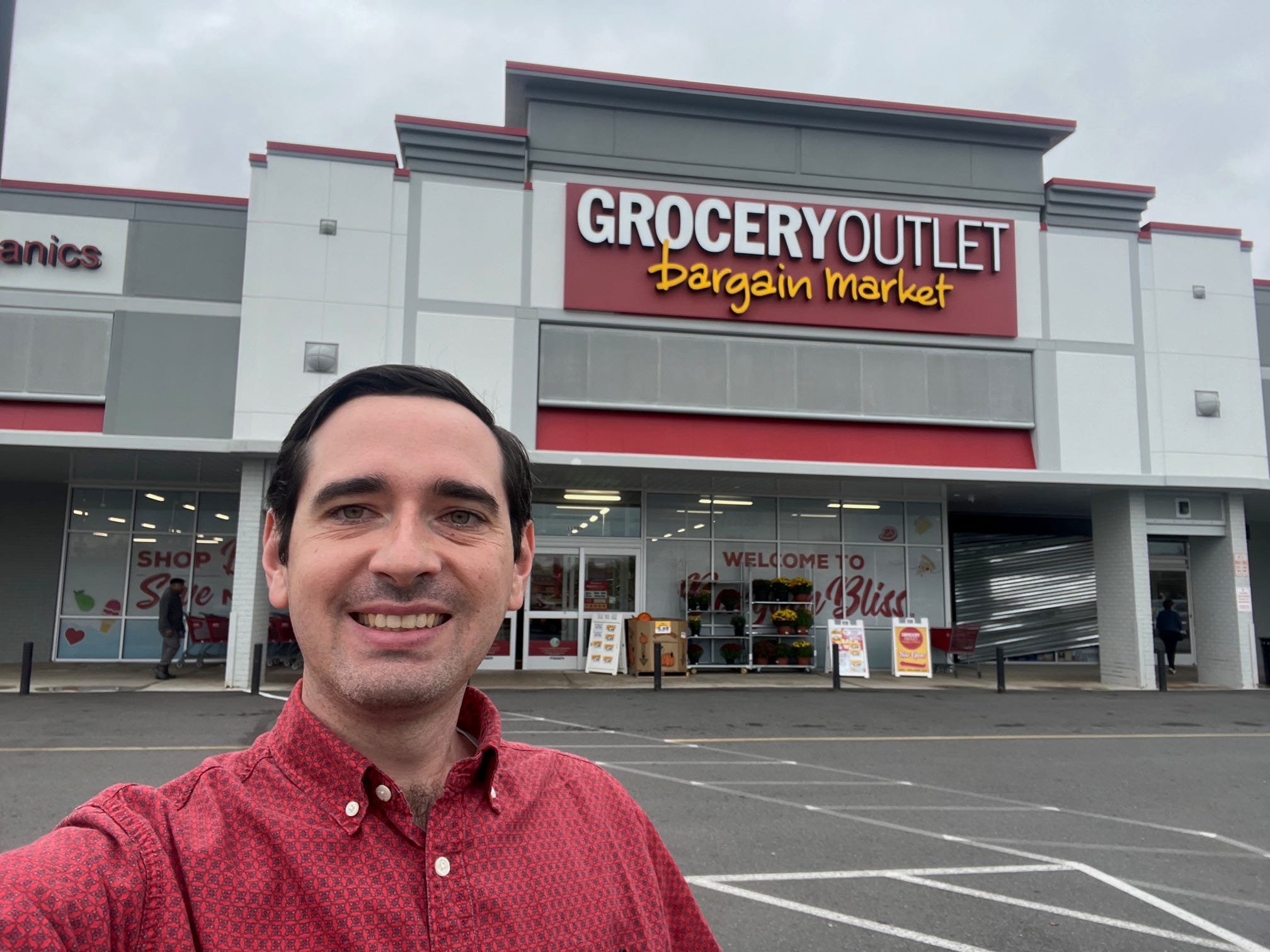 Business Insider reporter Alex Bitter stands in a red button-up shirt smiling outside of a Grocery Outlet supermarket parking lot. The sign above the entrance reads