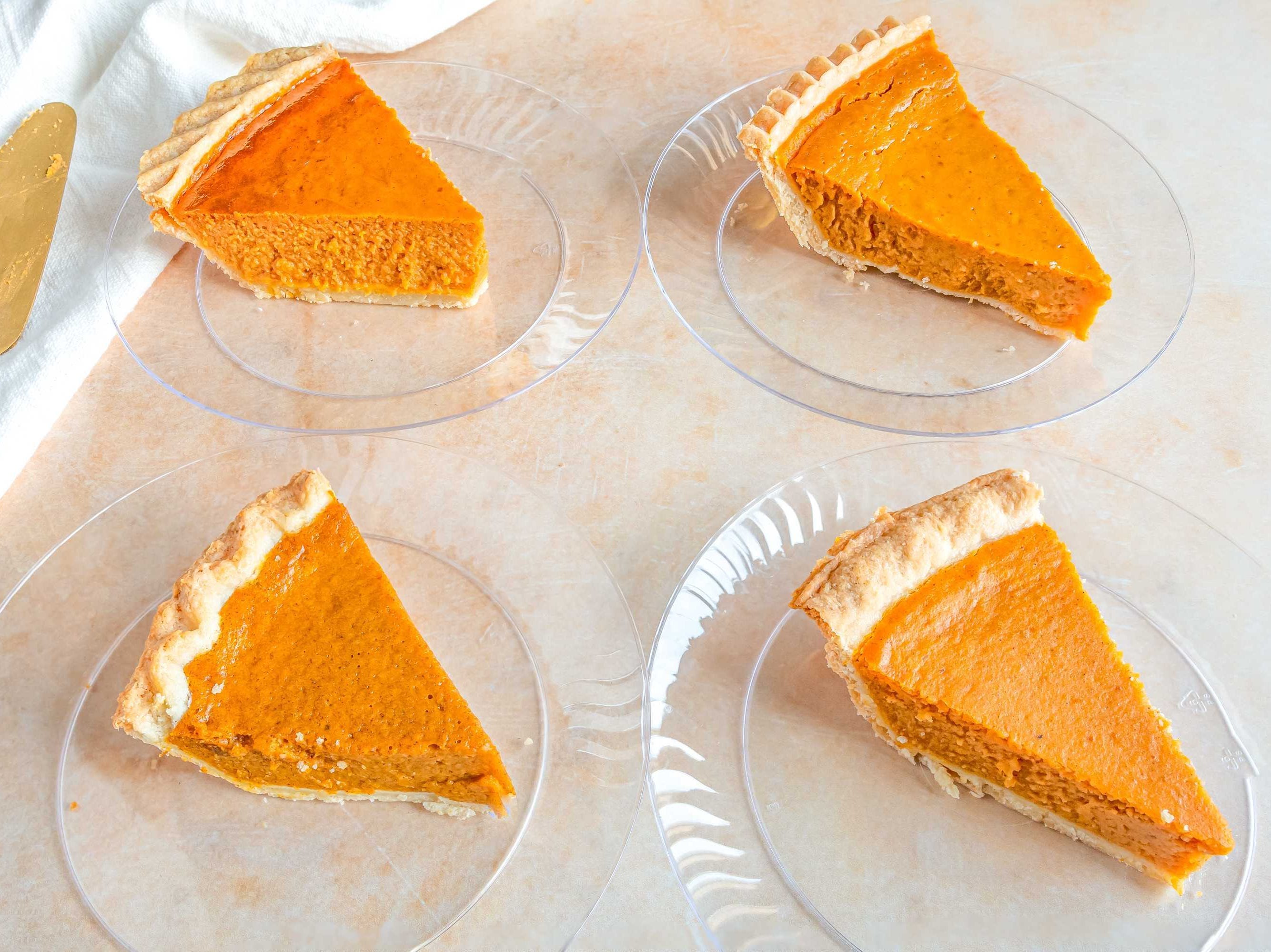 Four slices of pie on clear-glass plates, in rows of two. The plates sit on a light-brown table with a serving utensil to the left of them.