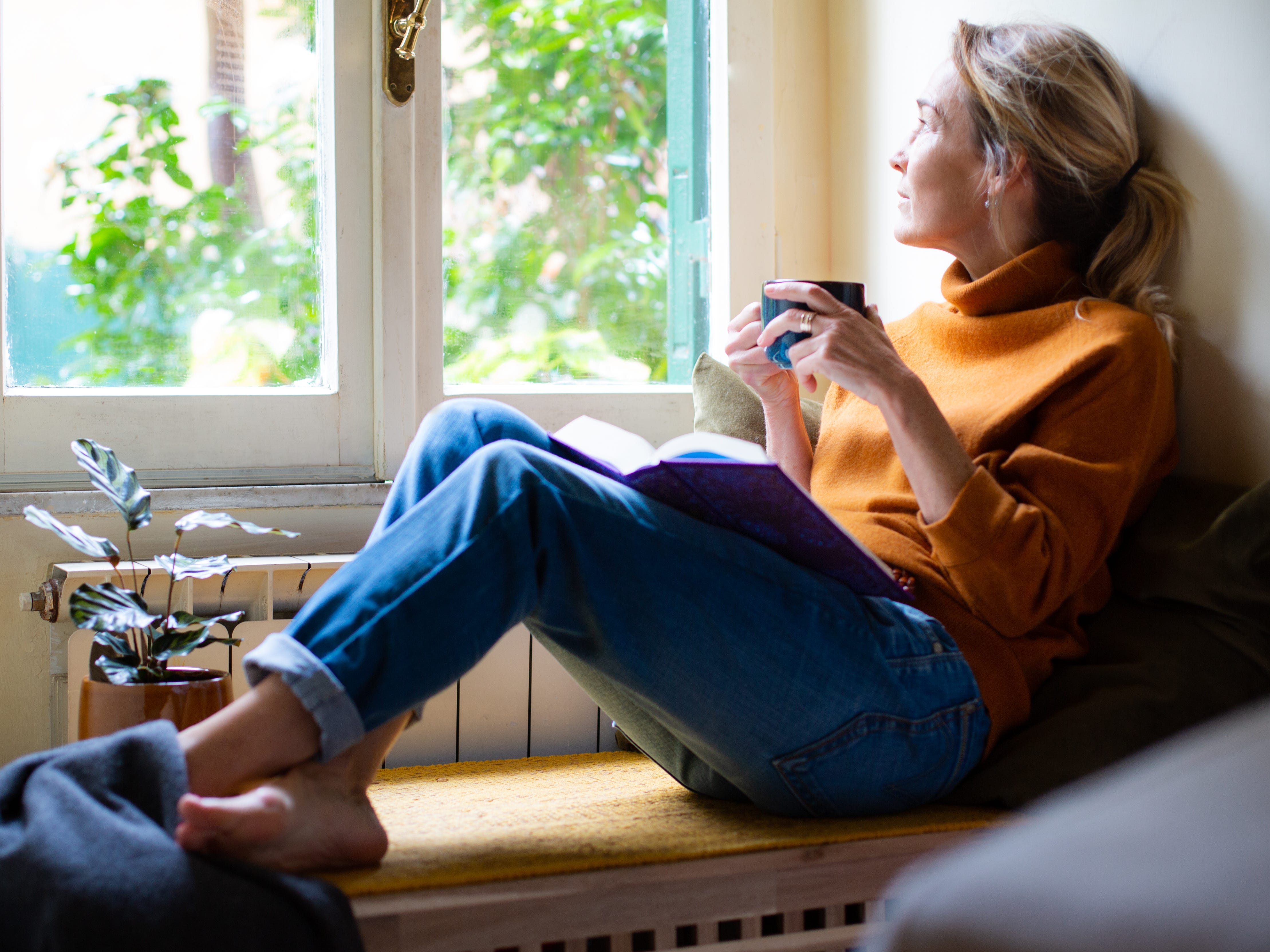 A woman drinking a cup of coffee and sitting on a windowsill while reading.