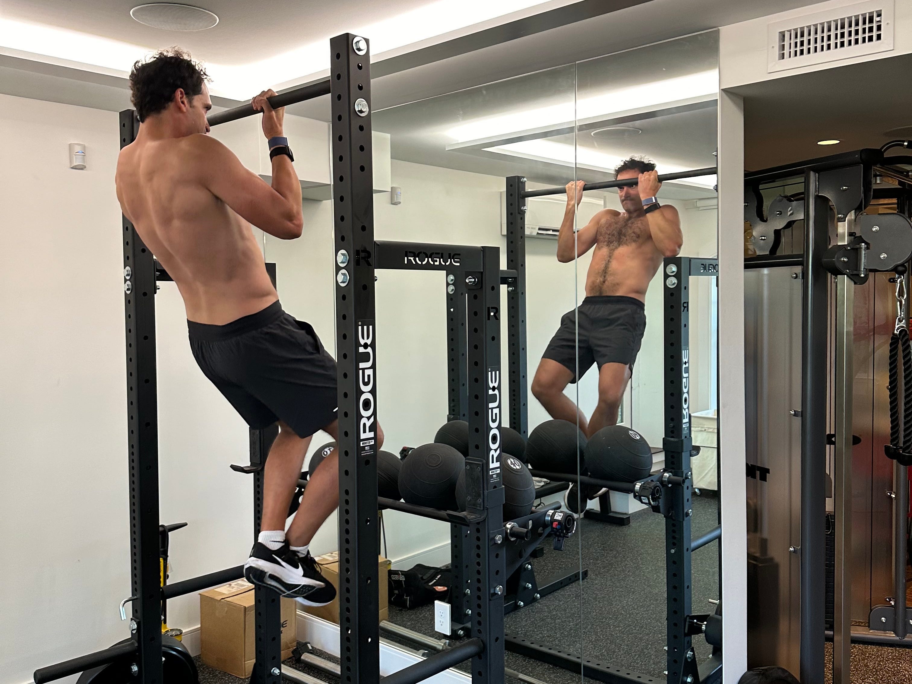 a man in black shorts and sneakers uses a pull-up bar in front of a mirror at the gym