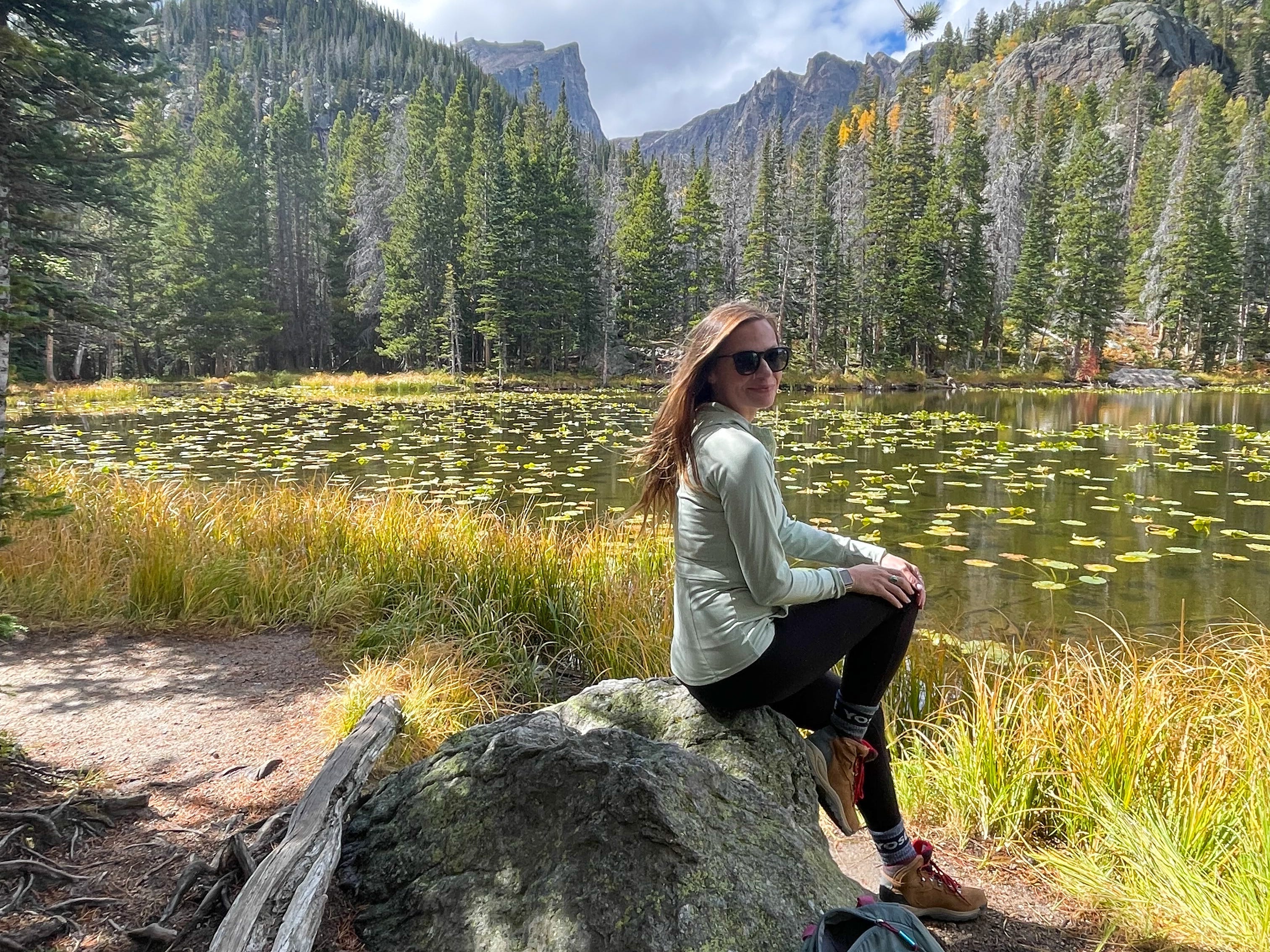 Emily sits on a rock with a lake and trees in the background in Rocky Mountain National Park.