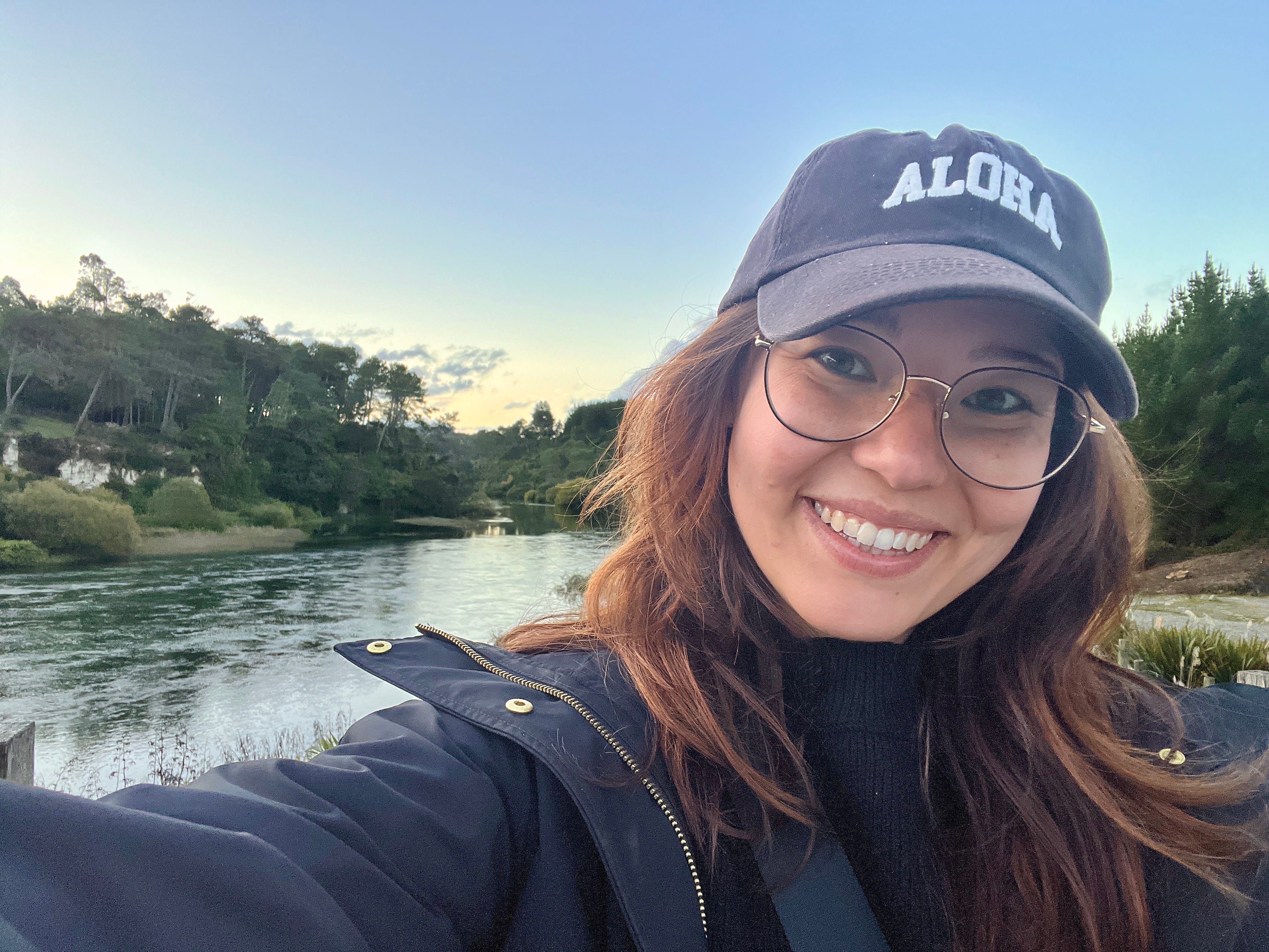 Ashley Probst taking a selfie with a body of water in the background. She's wearing glasses and a hat that says,