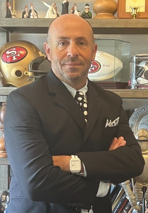 A man in a dark suit and polka dot tie stands with arms crossed in front of shelves filled with sports memorabilia