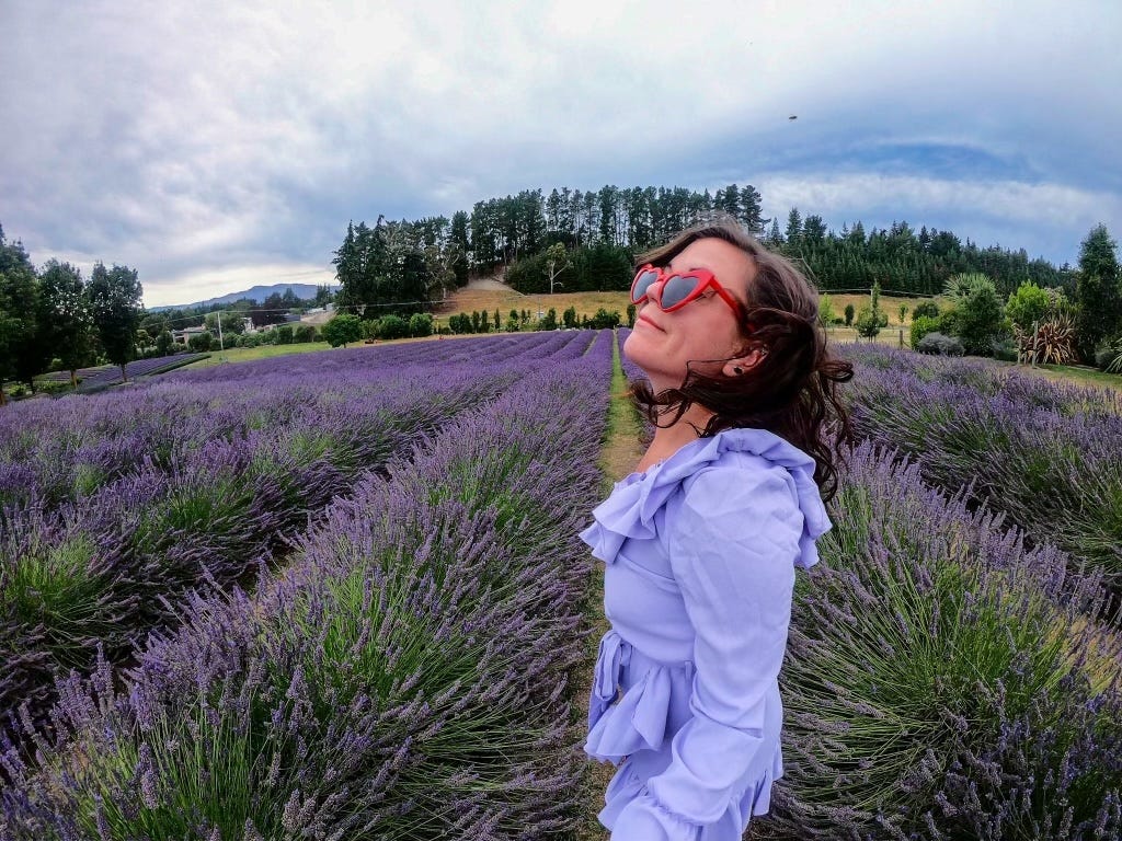 The author standing in fields of lavender in Wānaka, New Zealand. She's wearing a light-purple ruffled outfit with red, heart-shaped sunglasses.