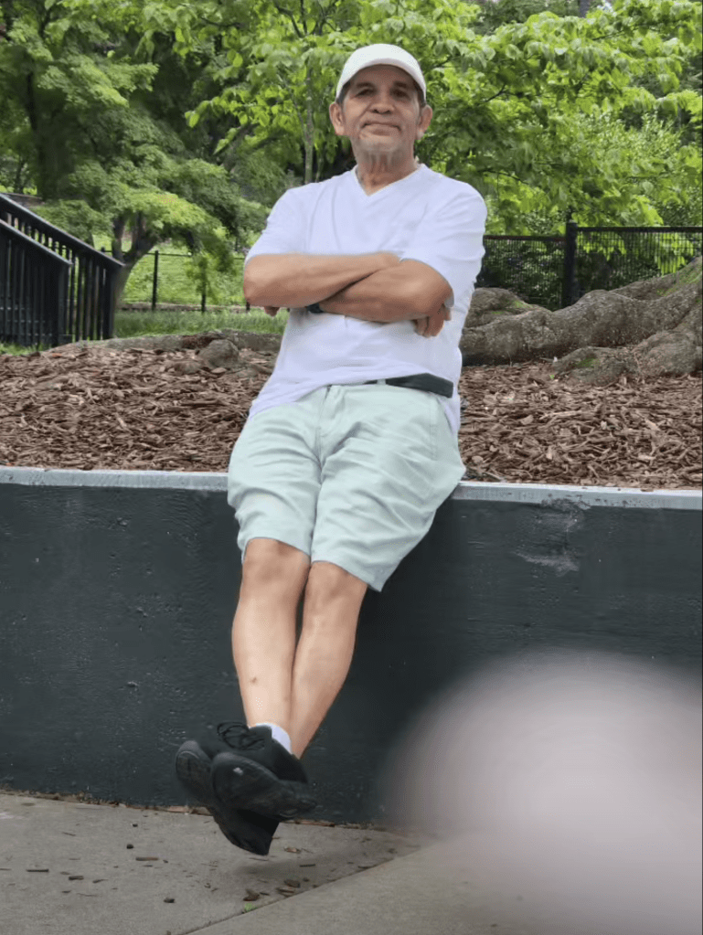Leobardo Hernandez, 64, wearing a white shirt and shorts, with arms crossed, leaning against a dark grey wall.