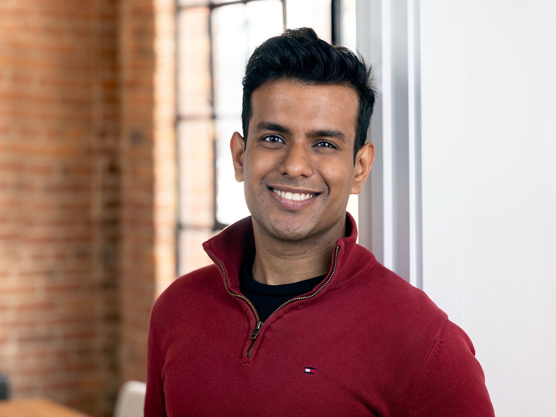 A young professional Indian man smiles standing in front of a window.