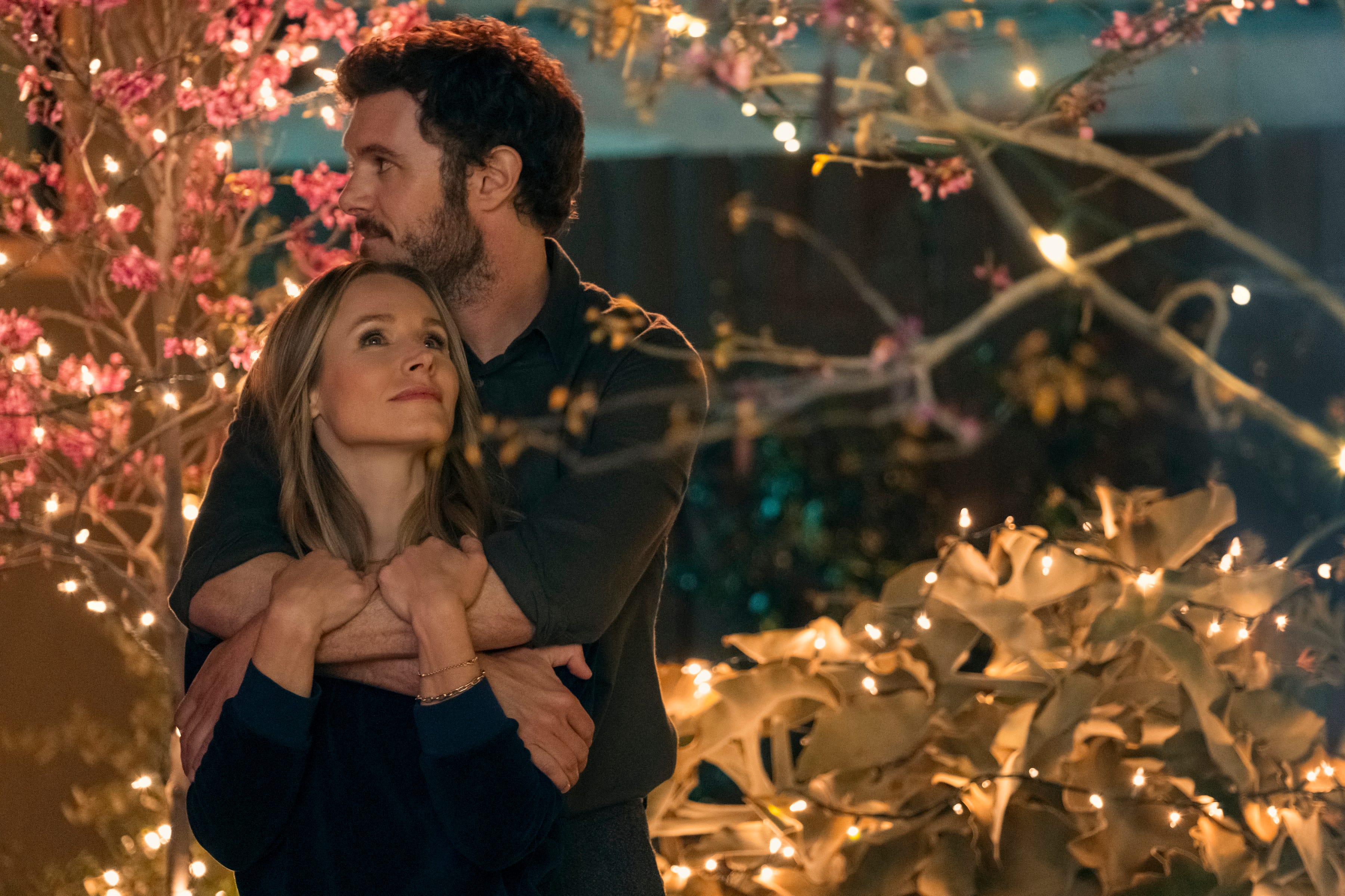 kristen bell and adam brody as joanne and noah in nobody wants this. they're standing outdoors in a string light lit garden, and noah has his arms around joanne and is resting his chin on her head