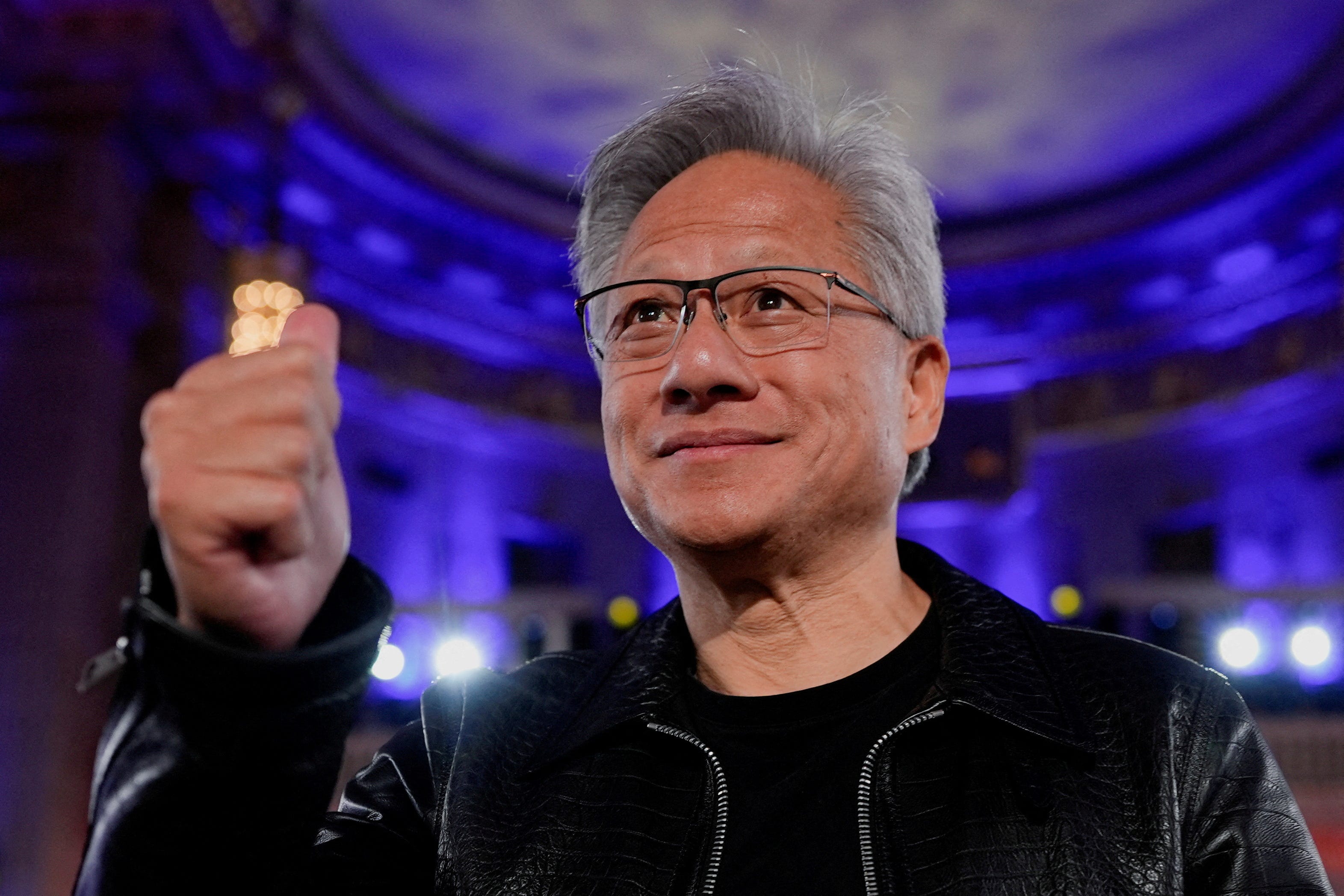 A grey-haired main, Nvidia CEO Jensen Huang, gives a thumbs up in a blue-lit room.
