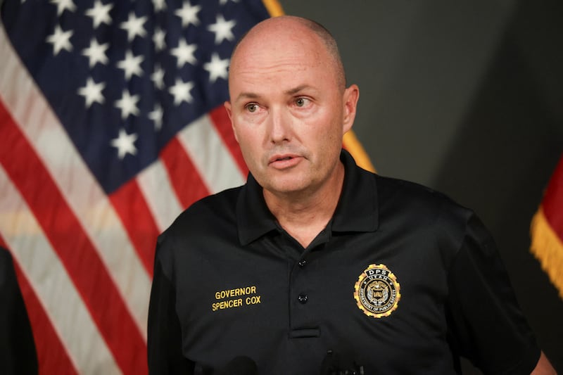 Utah Governor Spencer Cox speaks. He is wearing a black polo with his name on it and standing in front of a U.S. flag