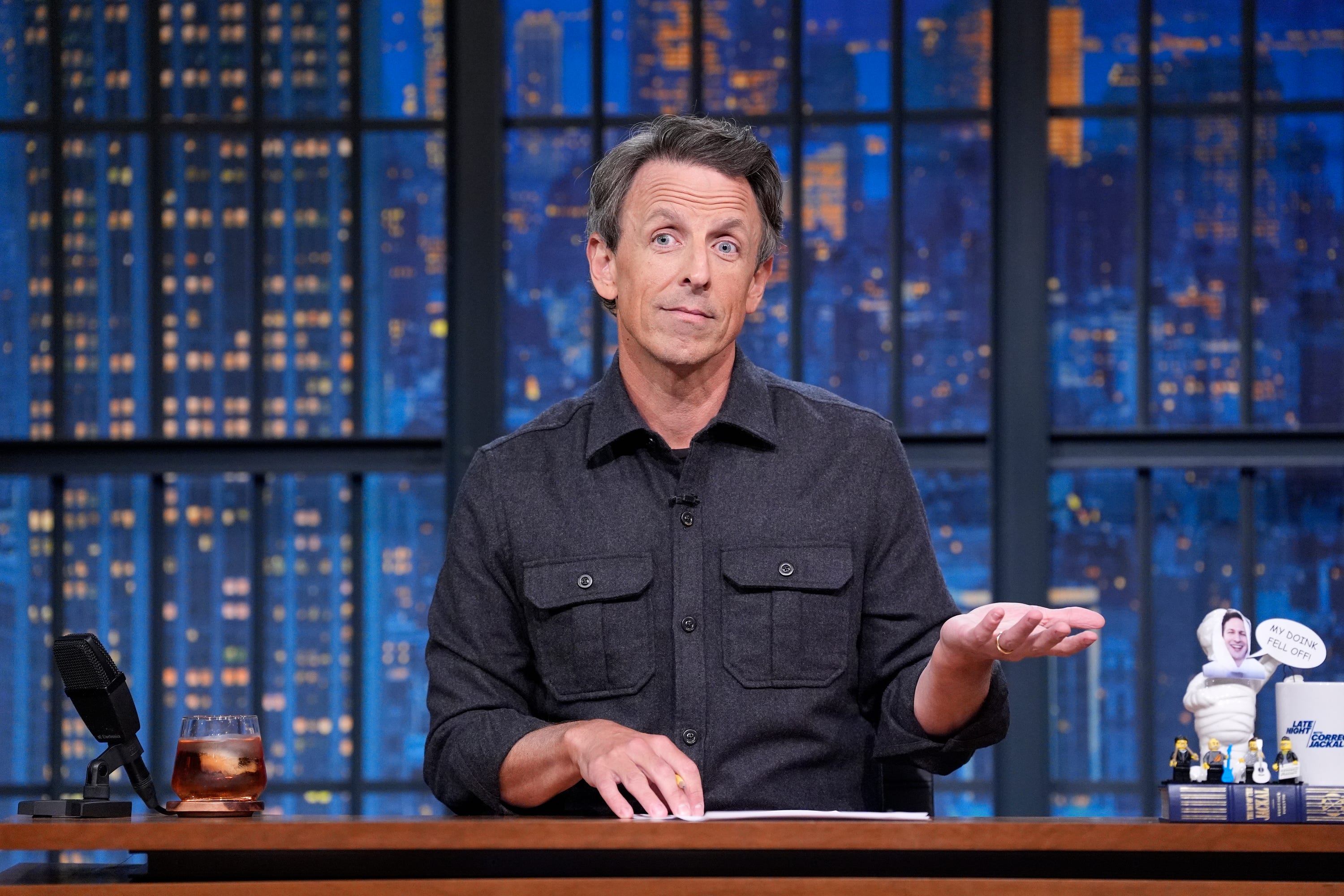 Seth Meyers is wearing a gray shirt while gesturing with his left hand.