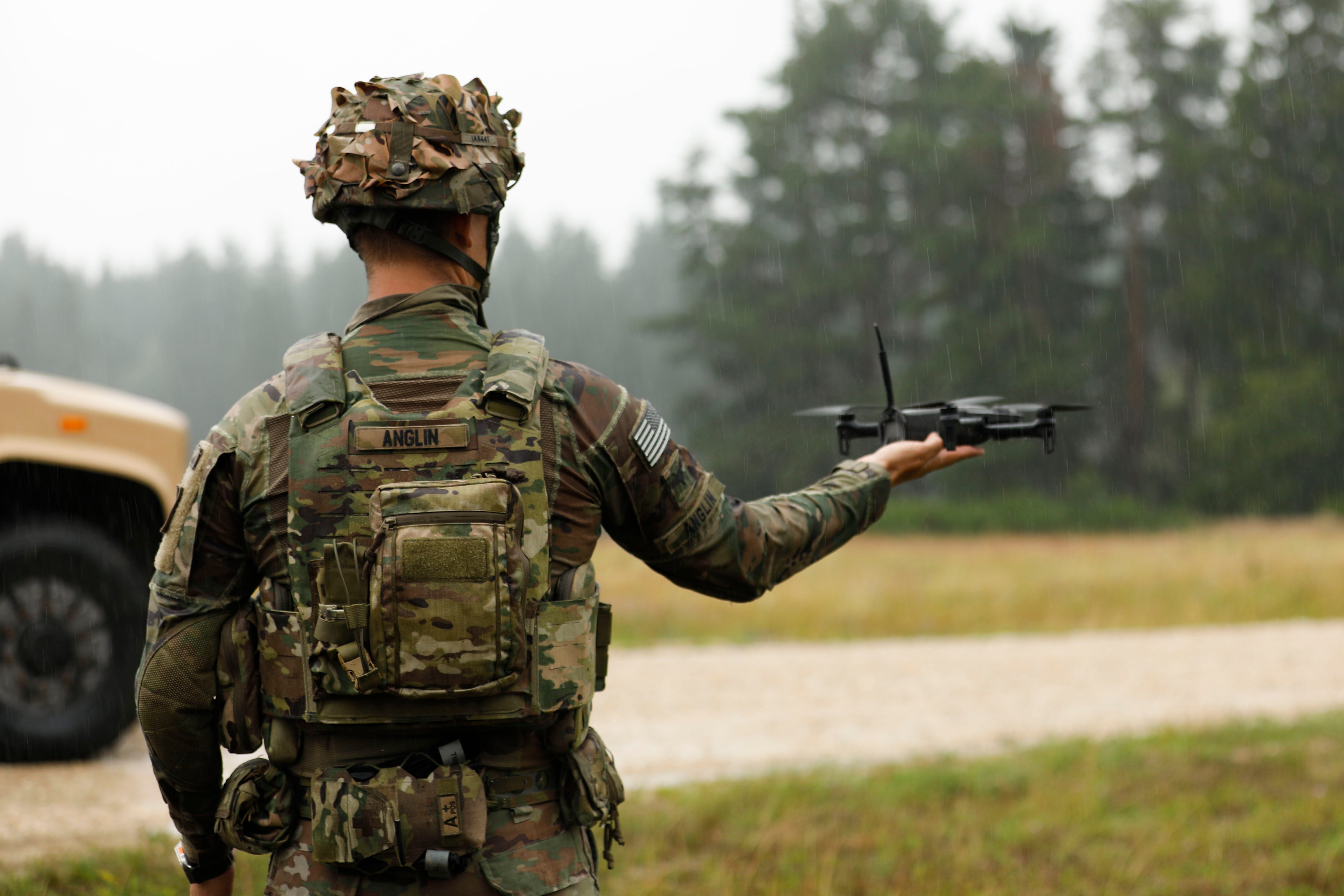 A soldier wearing camouflage stands with his back to the camera holding a small black drone in his right hand. In the background, a blurry road, trees and grass, and the front of a vehicle are seen in the rain.