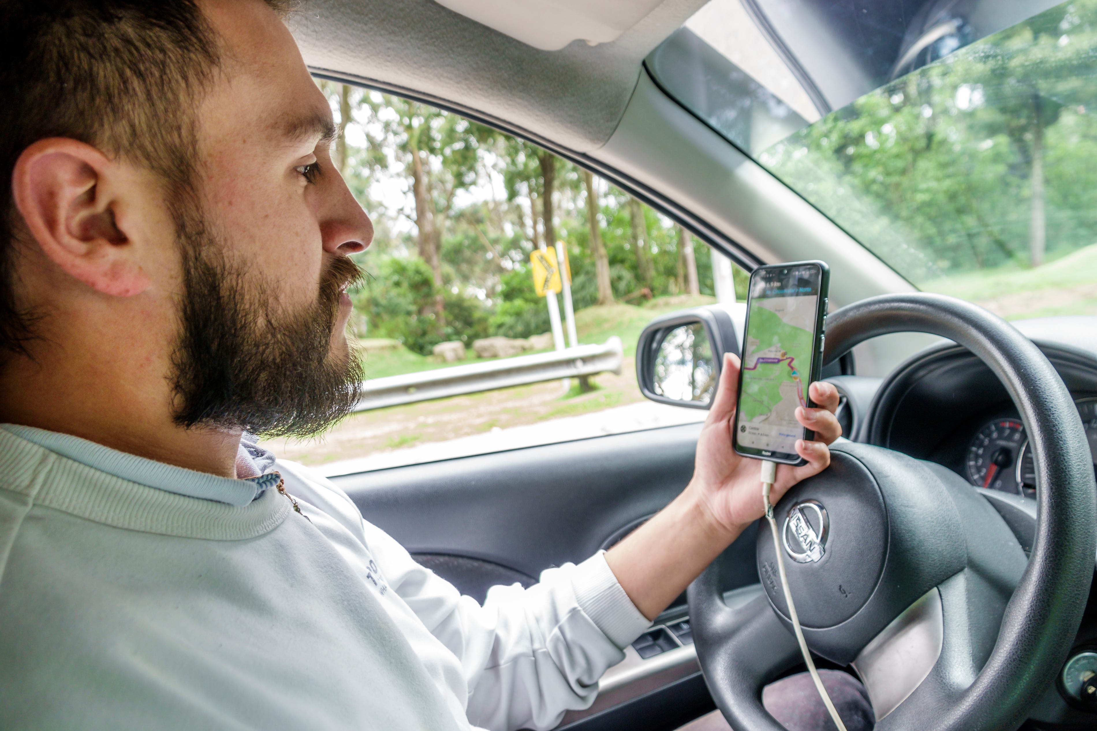 An Uber driver with a beard and a light-green shirt sits behind the wheel of a car and looks at a smartphone screen while it's plugged into his charging cable