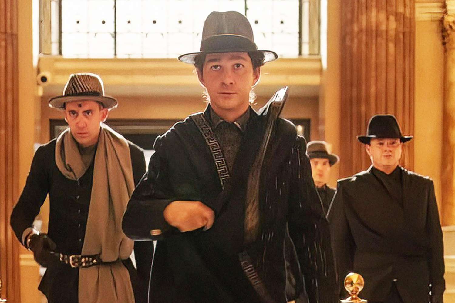 Shia LaBeouf wearing a hat and coat flanked by men in suits
