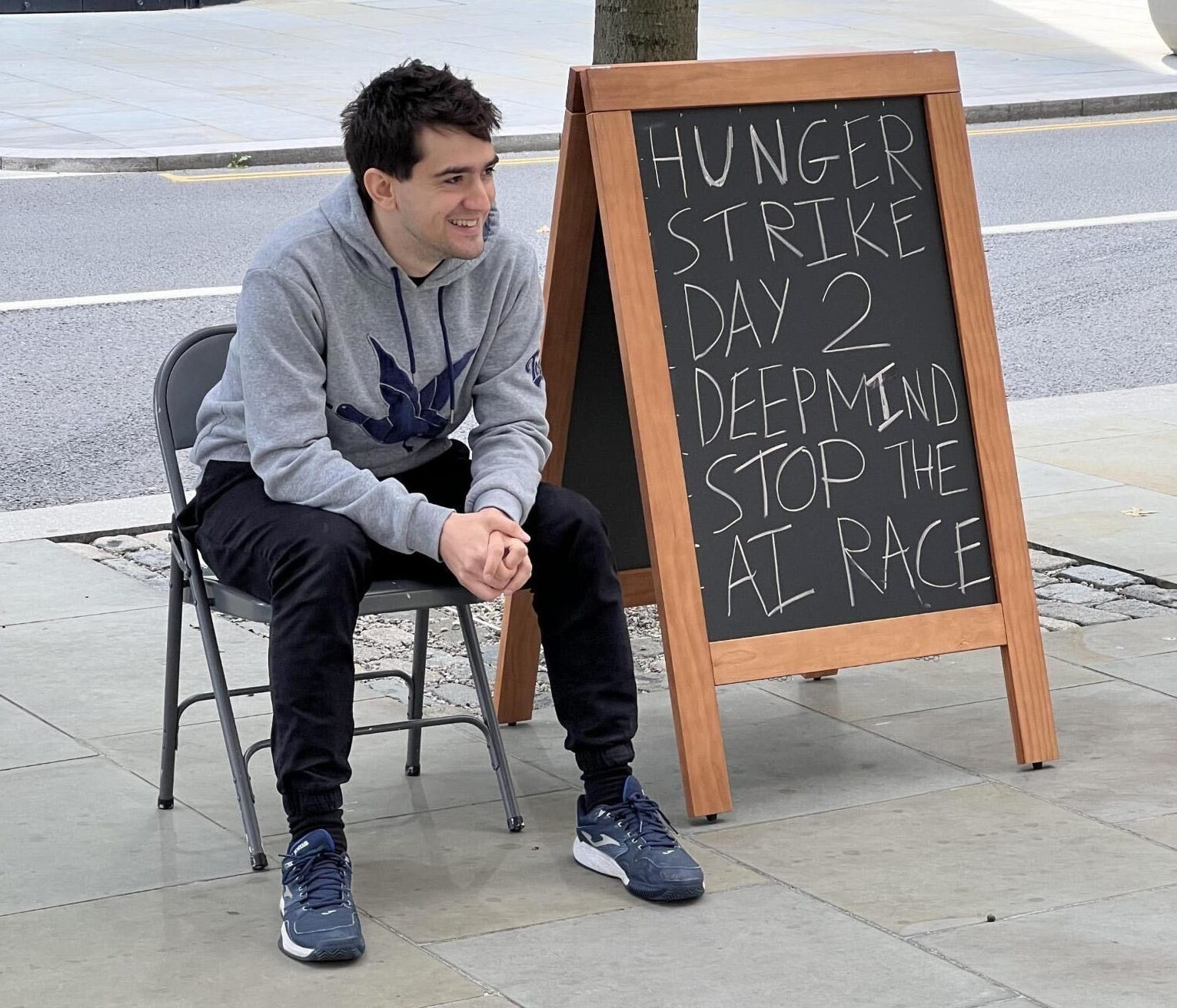 A man sits next to a sign that reads, "Hunger strike Day 2 Deepmind stop the AI race."