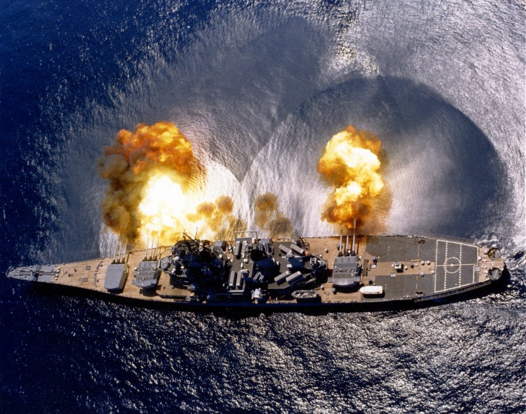The US Navy battleship USS Iowa seen from above firing guns while sailing in blue waters.
