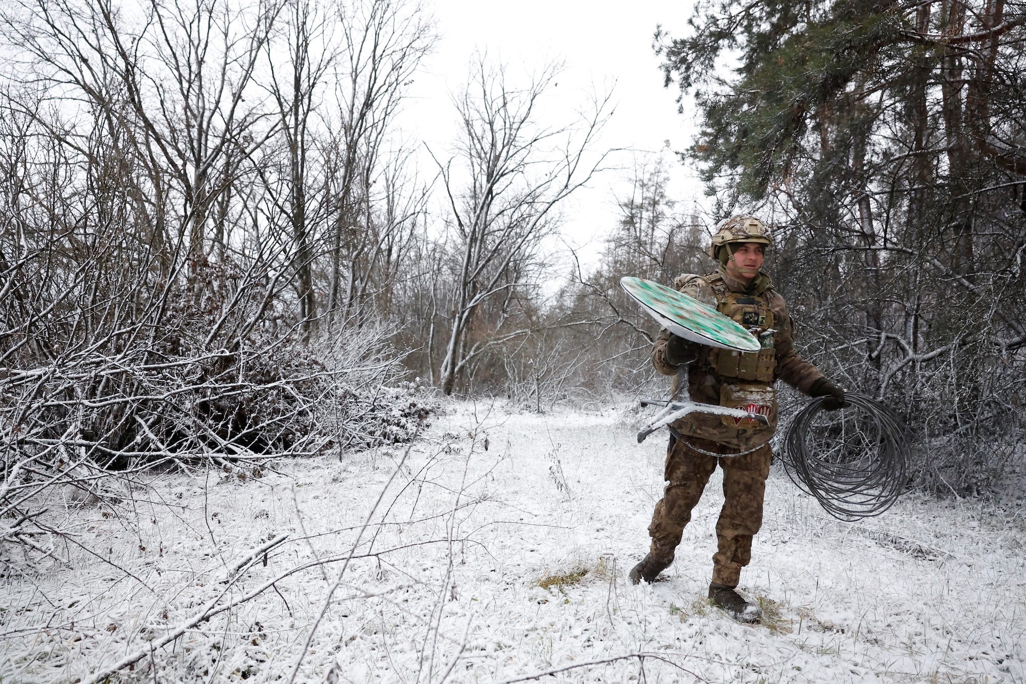 A man in camouflage gear holds a large green and white disk while standing in a snowy landscape