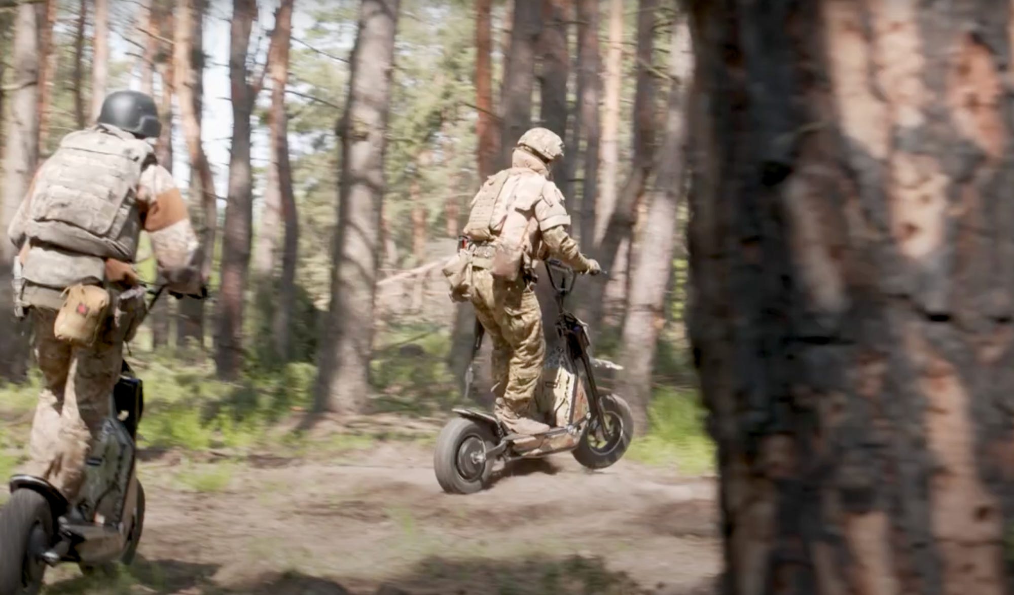 A still from video footage shows two people in camouflage gear riding large scooters through trees
