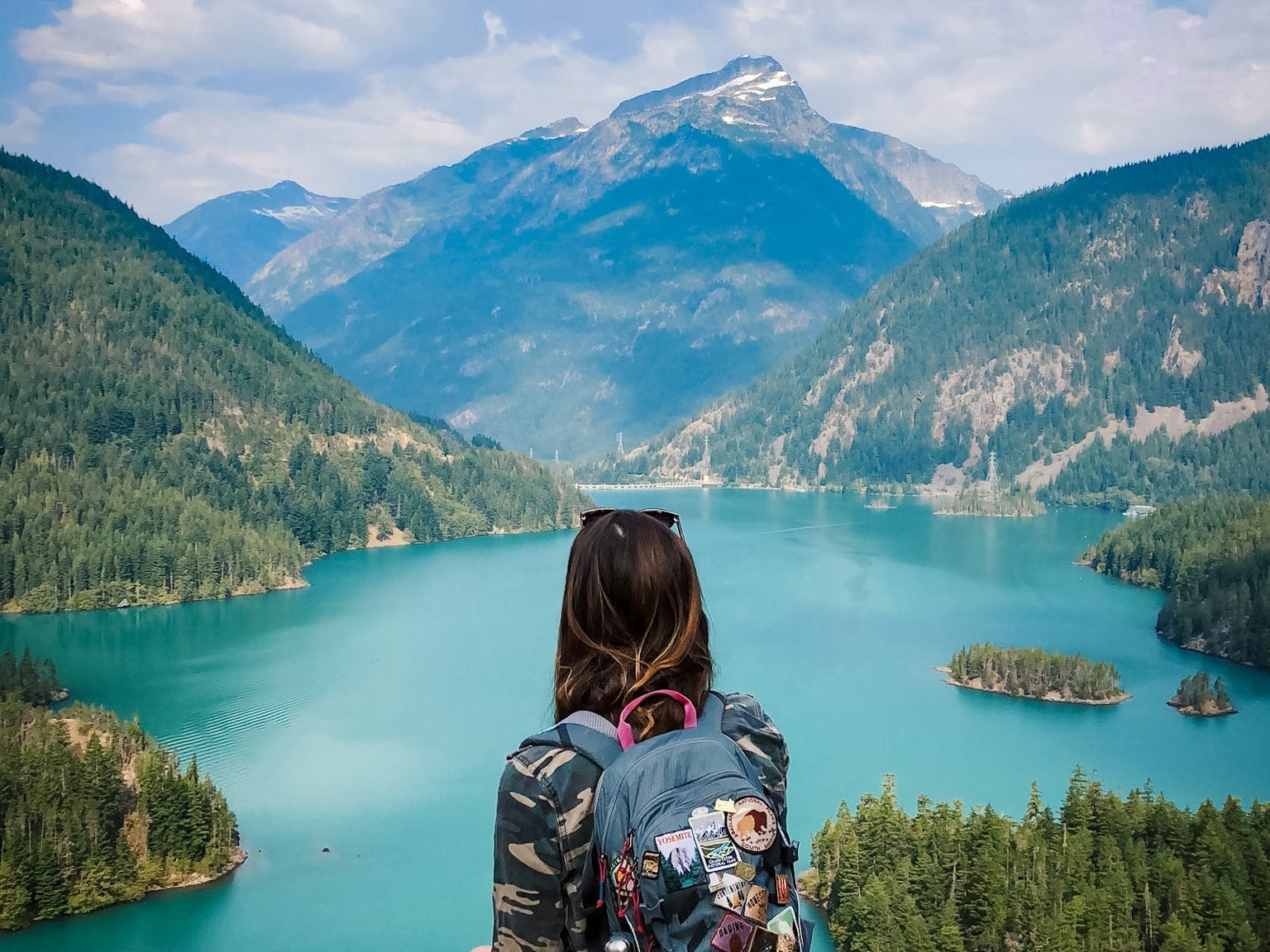 Emily, wearing a backpack covered with patches, looks out at a lake with blue waters and mountains covered in trees.